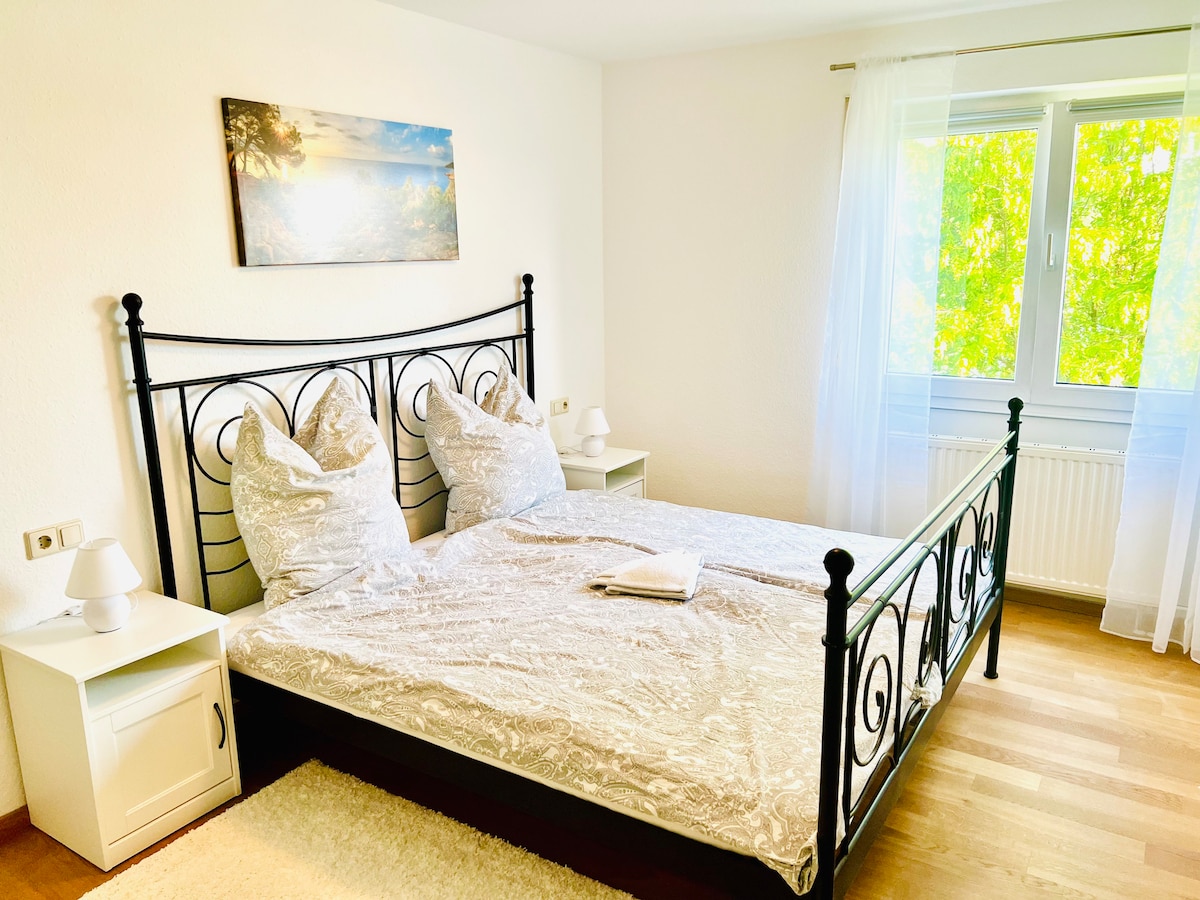 A bright bedroom features a stylish black wrought iron bed with a light-colored quilt. Two bedside tables with lamps are placed on each side of the bed. A large window allows natural light to illuminate the space, with a view of green foliage outside.