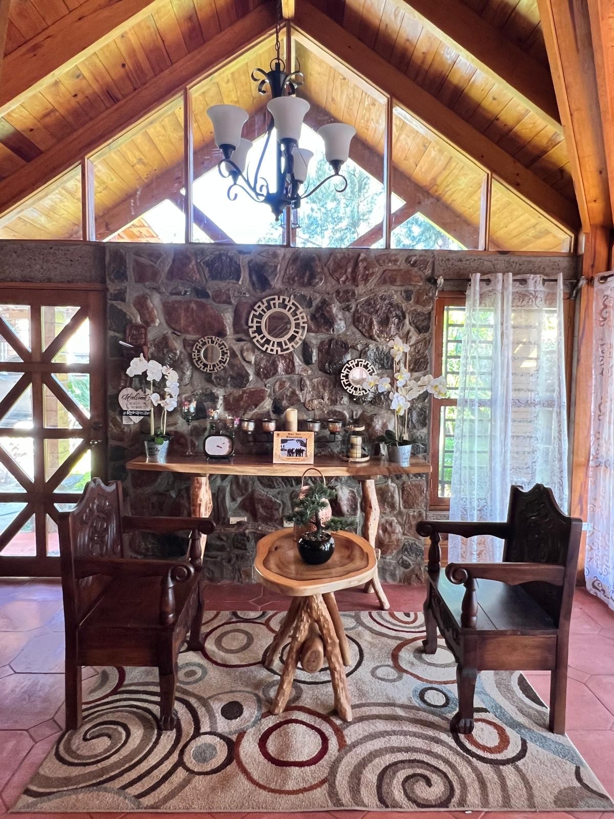 A welcoming entry area features a stone accent wall and large windows allowing natural light to fill the space. Two wooden chairs are positioned around a small table, complemented by decorative plants and wall art. A patterned rug adds texture to the floor.