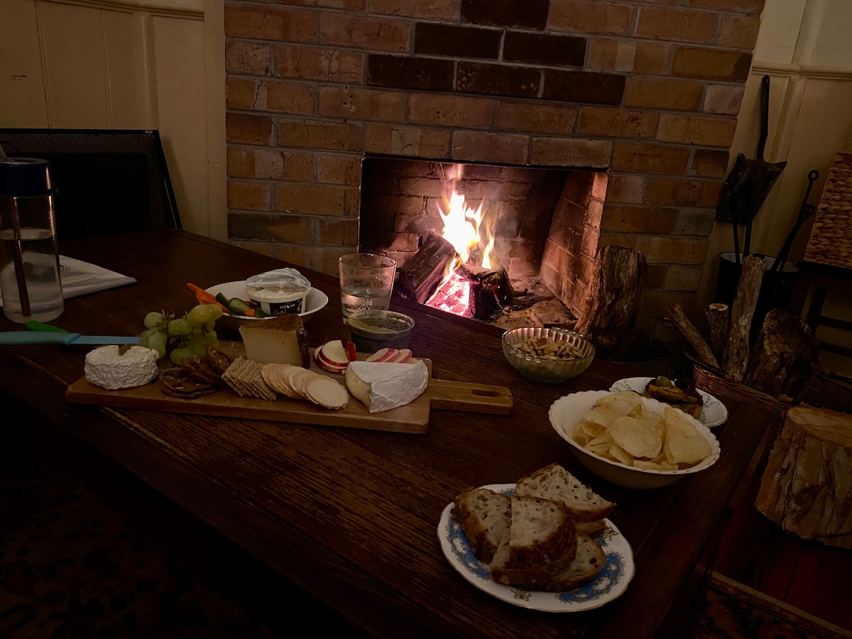 A wooden table is set with a variety of cheeses, crackers, and fresh fruits, arranged on a platter. A plate with chips is visible alongside slices of bread. A warm fire burns in a brick fireplace, providing a cozy ambiance.