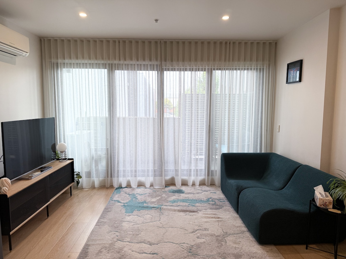 A spacious living area is filled with natural light, featuring large sheer curtains that softly filter the sunlight. A custom designer sofa sits invitingly, and a light-colored area rug adds a cozy touch. A television is positioned on a media console, creating a relaxed atmosphere.