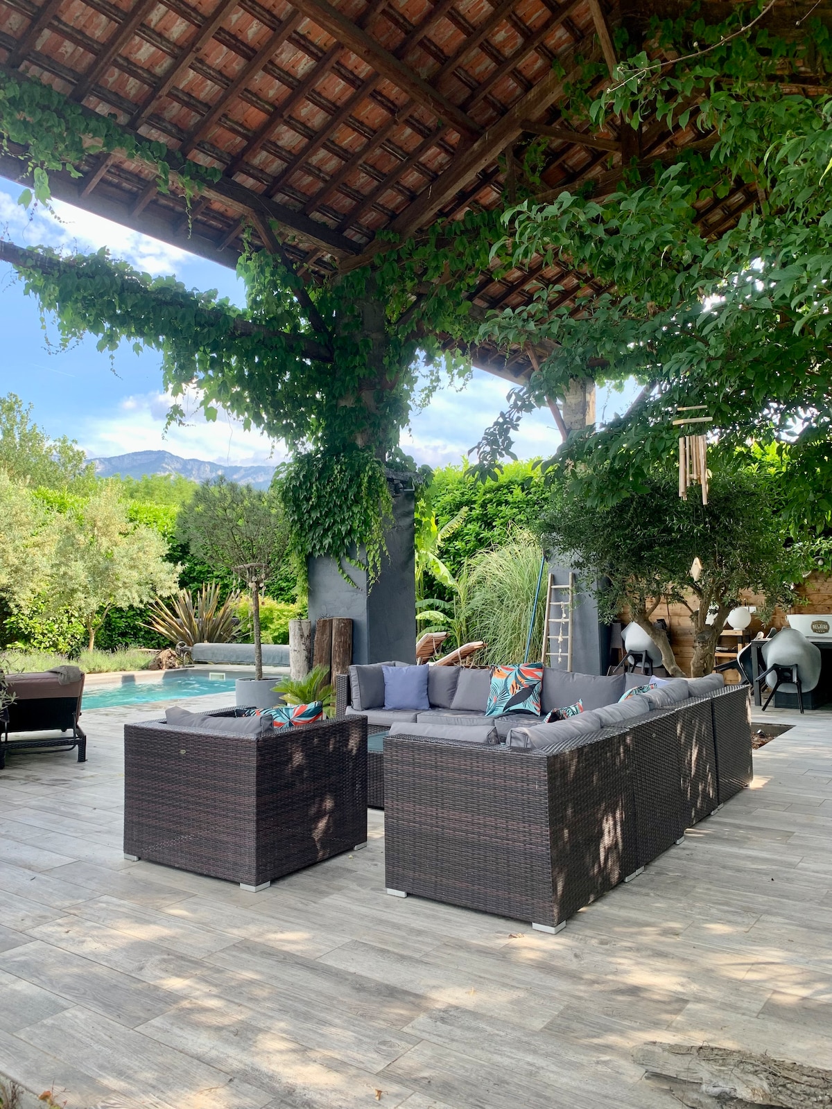 An outdoor lounge area features a set of woven sectional sofas positioned under a rustic wooden roof, surrounded by lush greenery. A spacious swimming pool is visible in the background, complemented by various plants and trees creating a serene ambience.