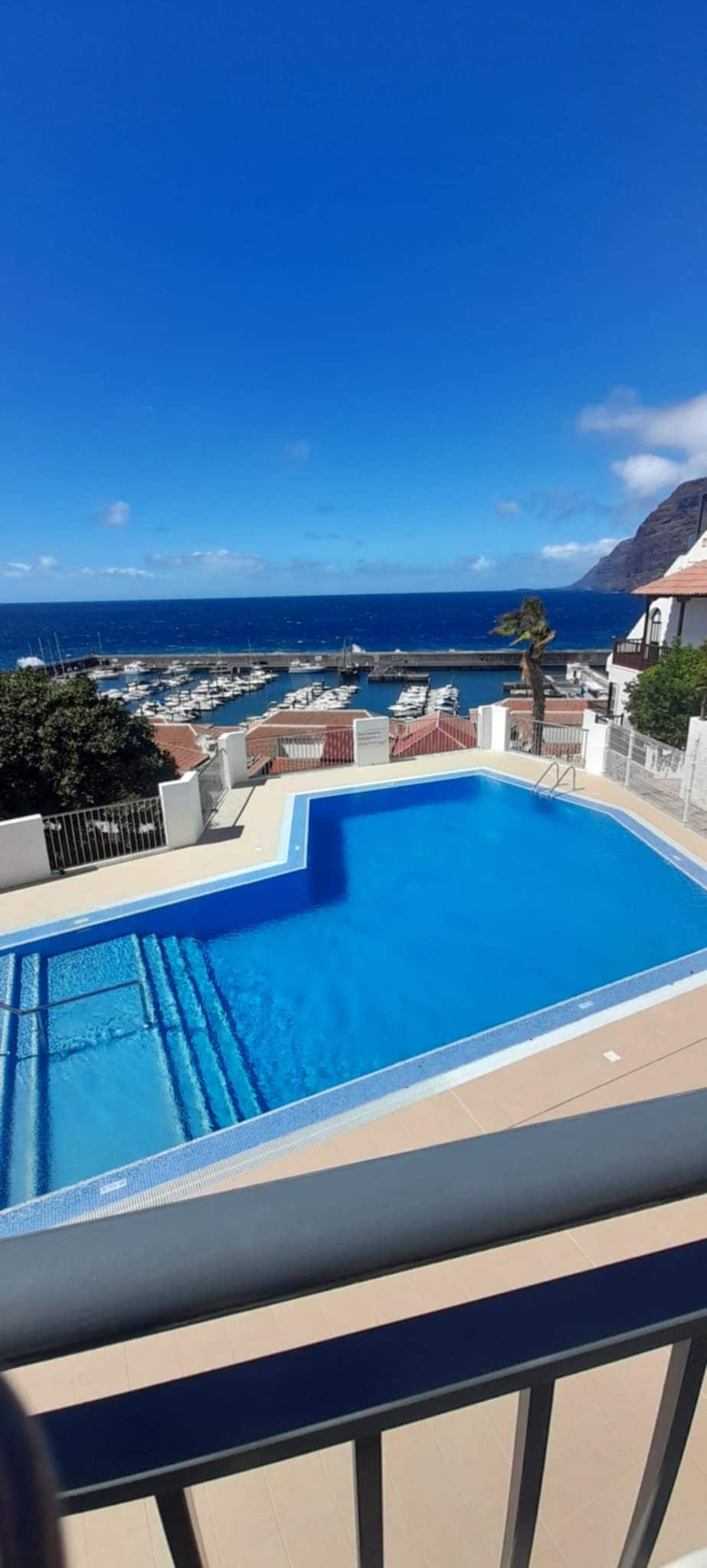 A clear blue pool is visible, surrounded by sun loungers and palm trees. The ocean can be seen in the background, with boats docked at a nearby marina. The scene is framed by a bright sky with scattered clouds, highlighting the tranquil setting.