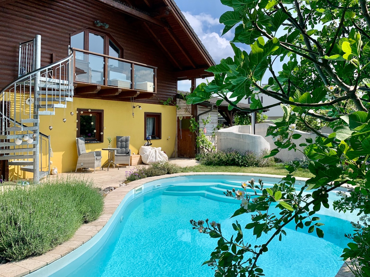 A bright, inviting outdoor space features a refreshing pool surrounded by lush greenery and stone paving. A staircase leads to a balcony with seating, while the yellow exterior of the house adds a cheerful touch. The area is framed by vibrant plants, enhancing the serene setting.