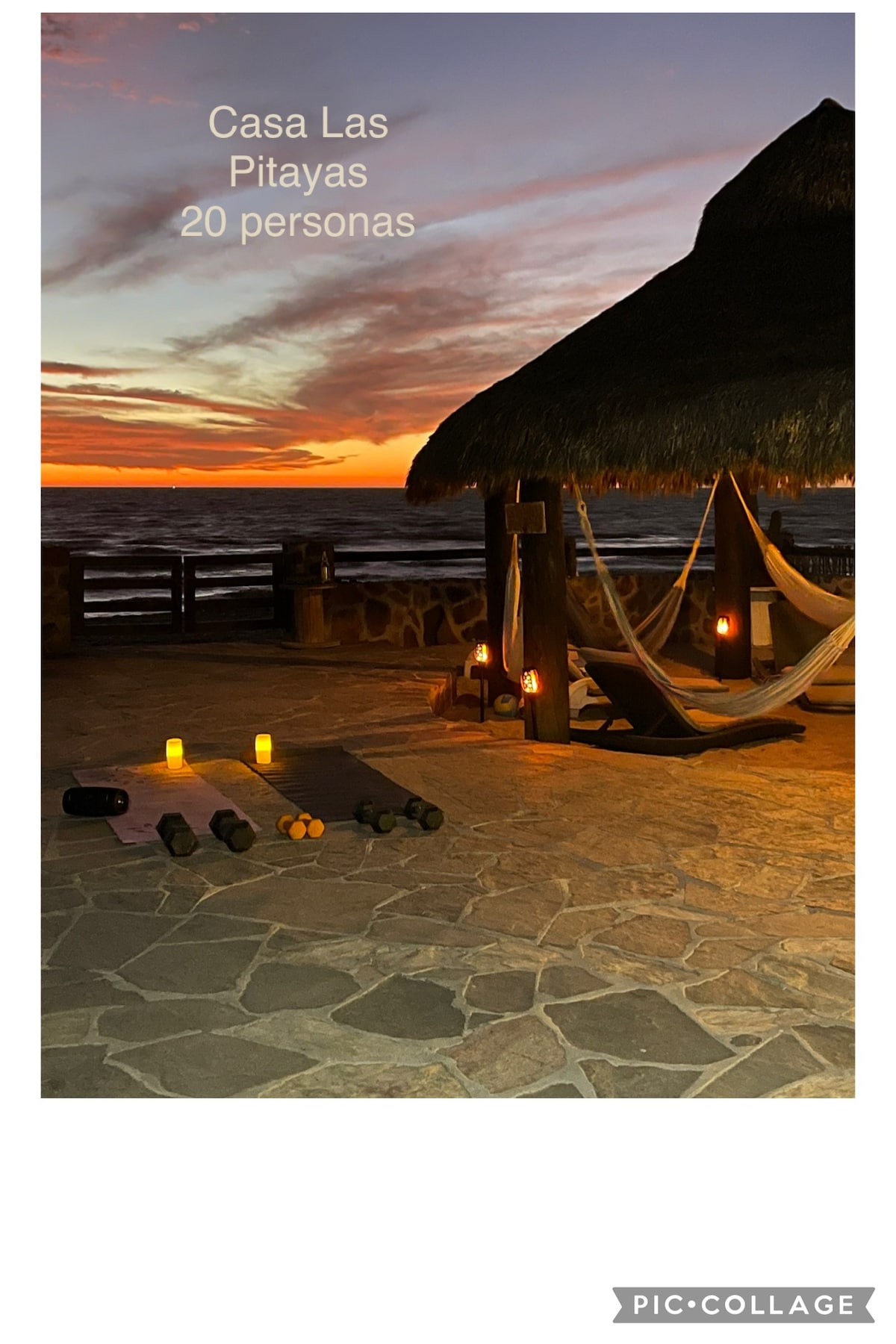 An outdoor seating area is highlighted by the warm glow of candles, surrounded by stone flooring. A traditional thatched roof provides shade over comfortable hammocks. The sunset casts vibrant colors across the sky, creating a serene ambiance near the water.