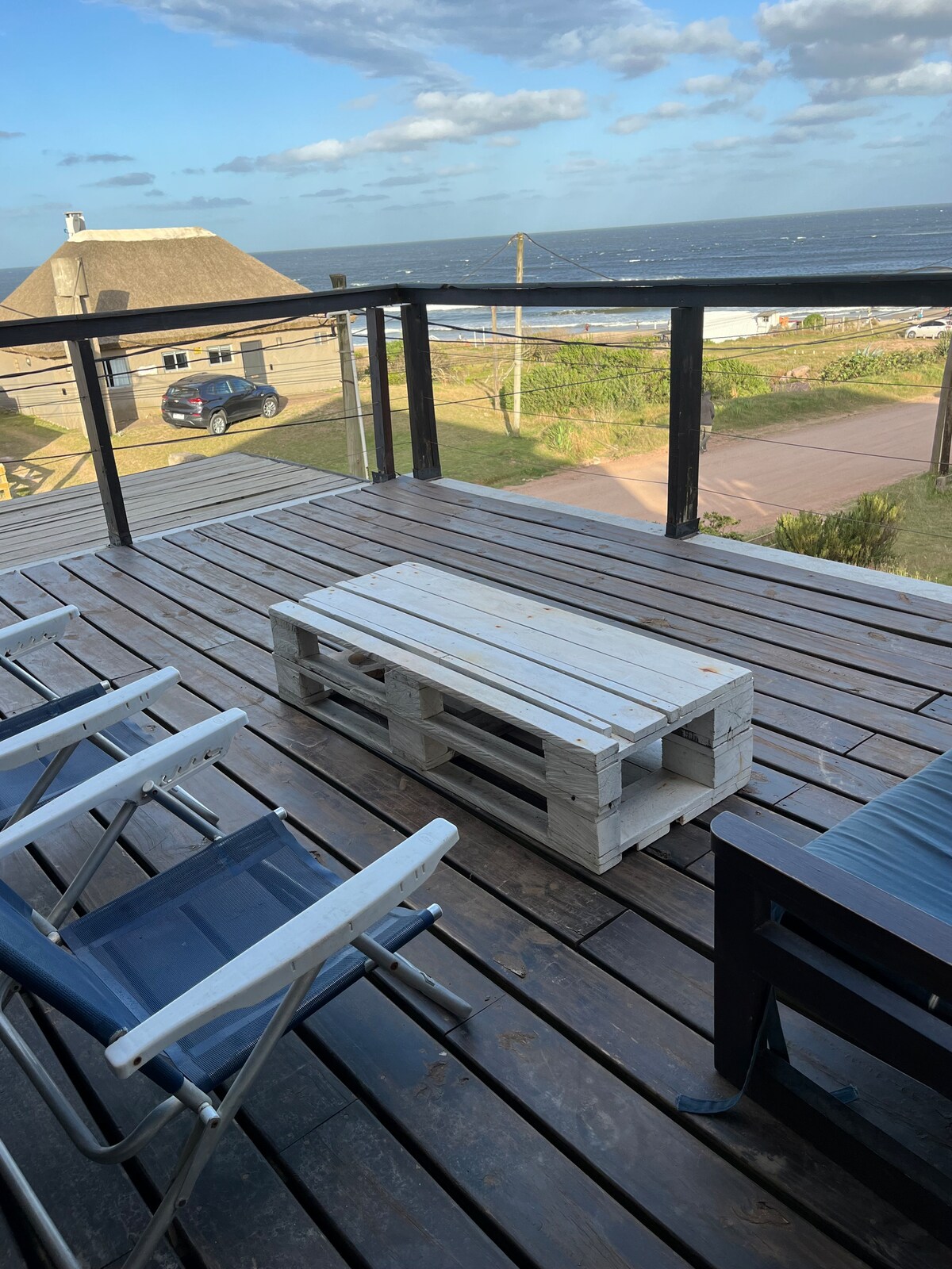 An outdoor terrace area features a wooden deck with a low coffee table made from pallets. Two folding lounge chairs are positioned nearby, offering views of the sea and the surrounding landscape. Accessible glass railings ensure a clear sightline to the ocean beyond.