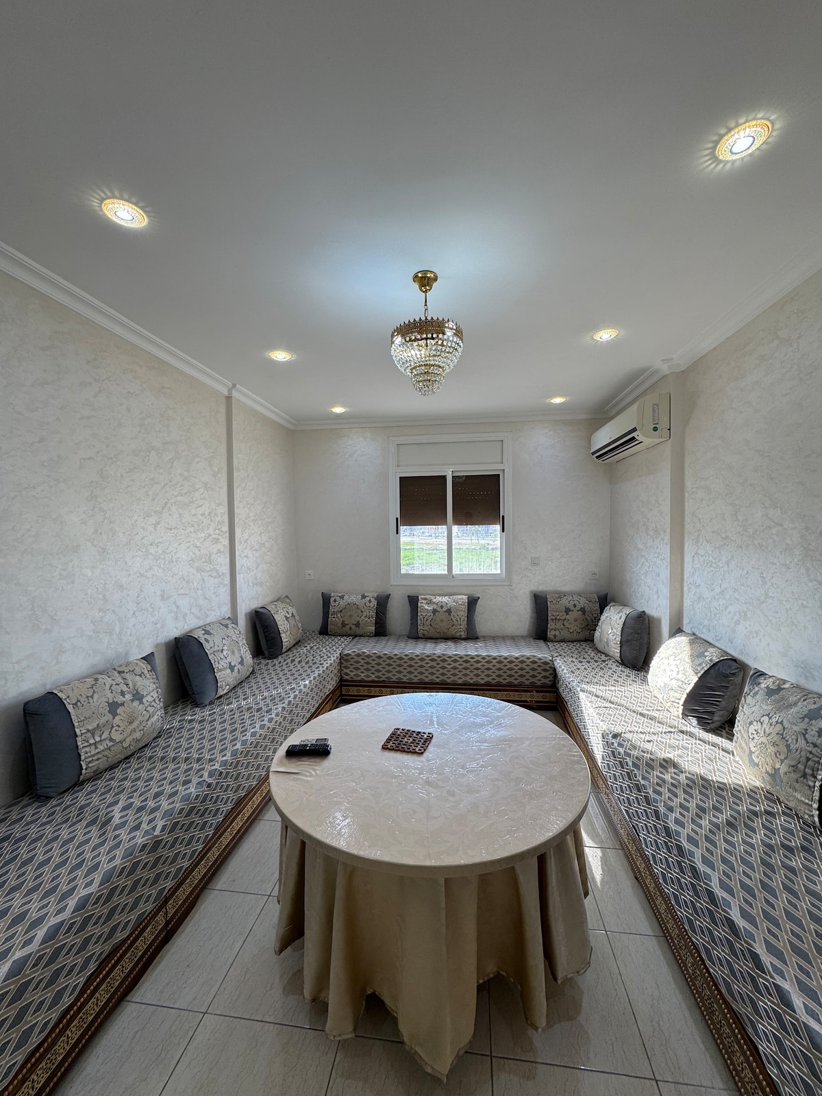 A comfortable seating area features a large circular table at the center, surrounded by cushioned benches with decorative pillows. A chandelier hangs from the ceiling, and natural light enters through a window, enhancing the serene space.