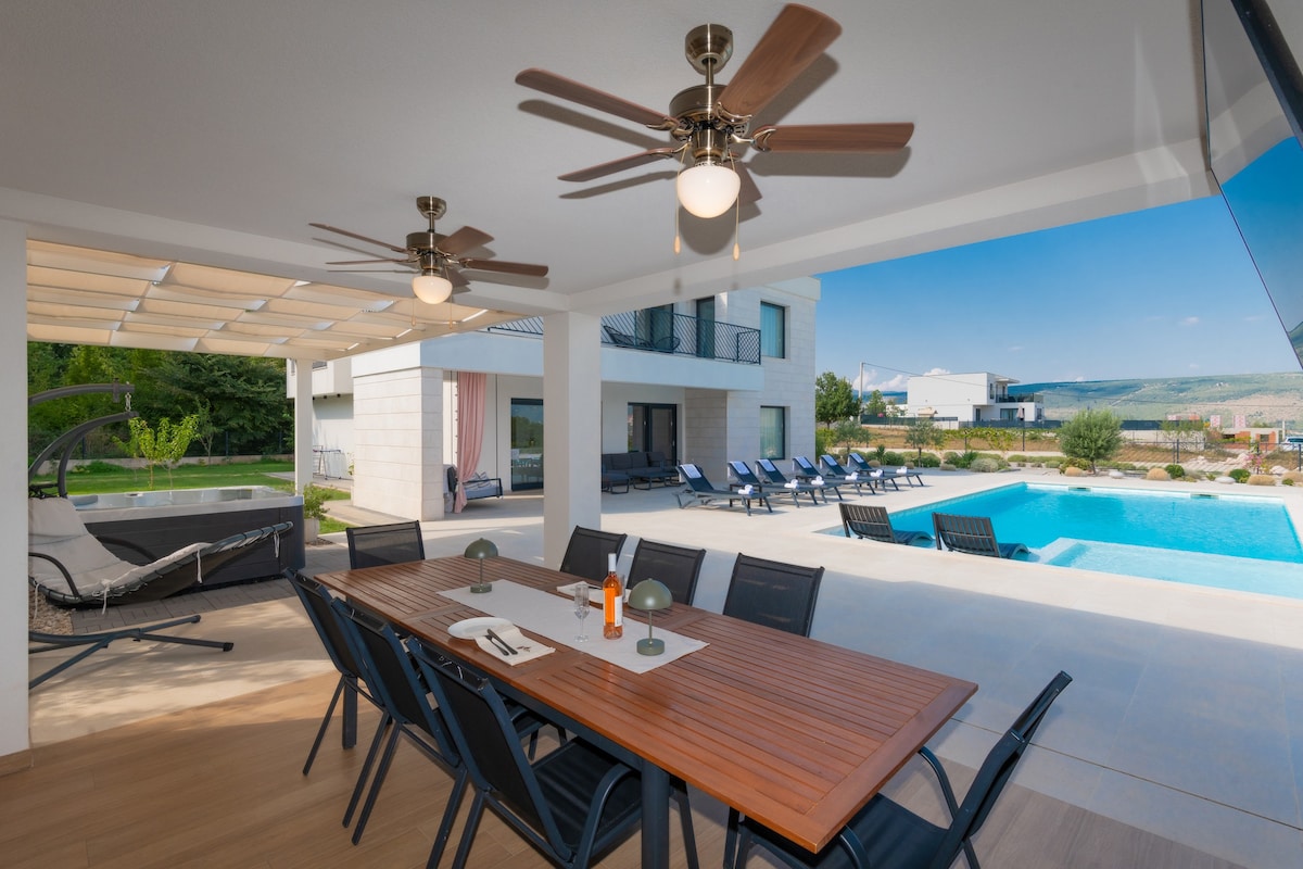 A covered outdoor seating area features a long wooden table surrounded by black chairs, ideal for dining. Ceiling fans enhance airflow, while a spacious pool and sun loungers are visible in the background, offering a relaxed atmosphere. Lush greenery frames the scene.