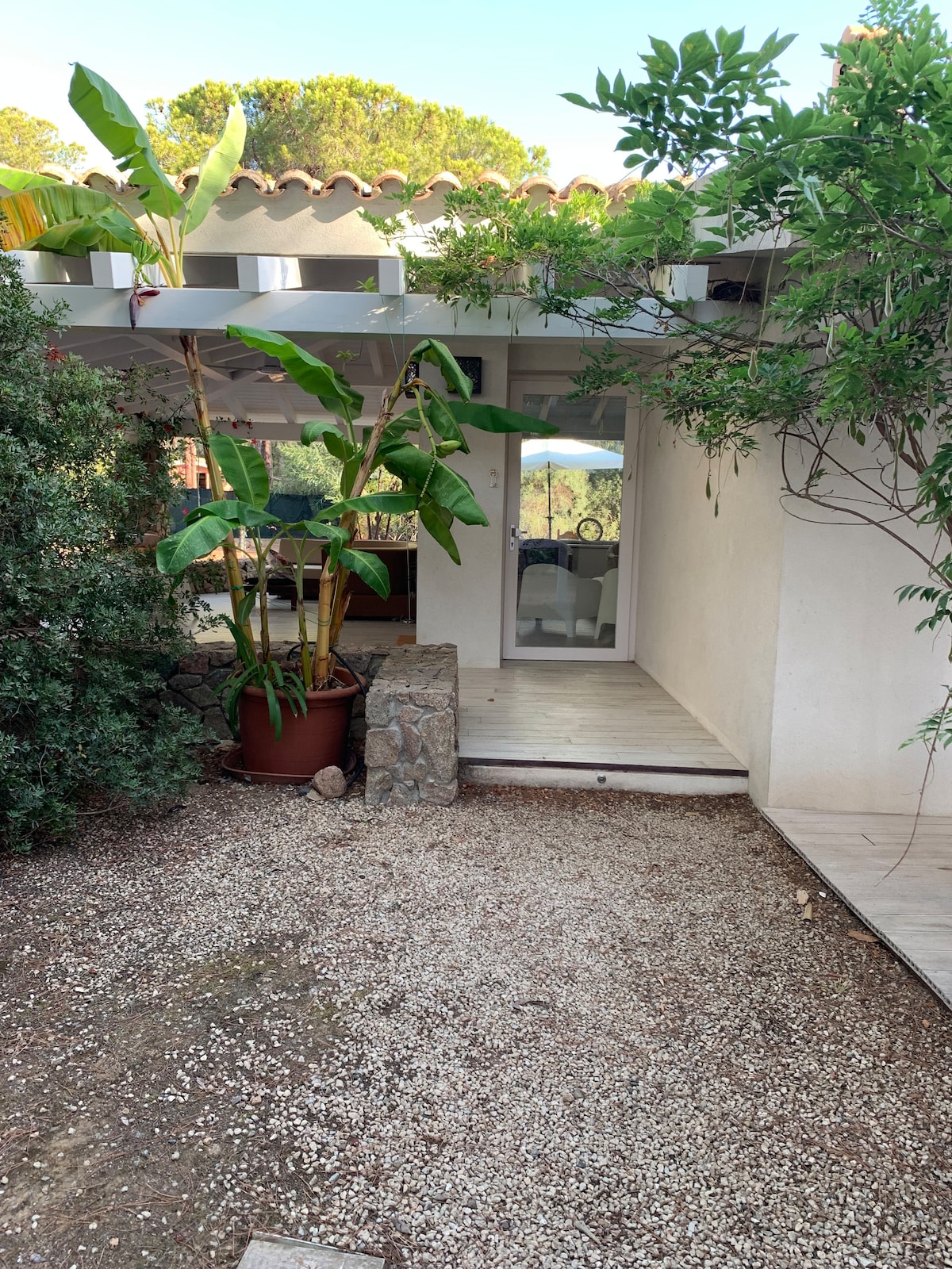 A pathway leads to a light-colored entrance, framed by lush greenery and tall potted plants. The area is accented with gravel and stone features, providing a natural, inviting ambiance. The structure blends with the surrounding garden, hinting at a tranquil retreat.