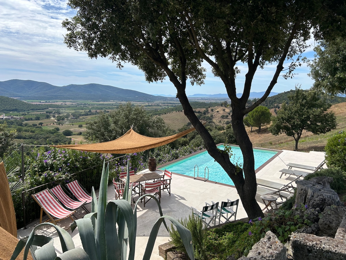 A private outdoor area features a swimming pool surrounded by sun loungers and a shaded table setting. Green hills and valleys are visible in the background, complemented by a clear sky. A large tree provides additional shade, enhancing the serene landscape.