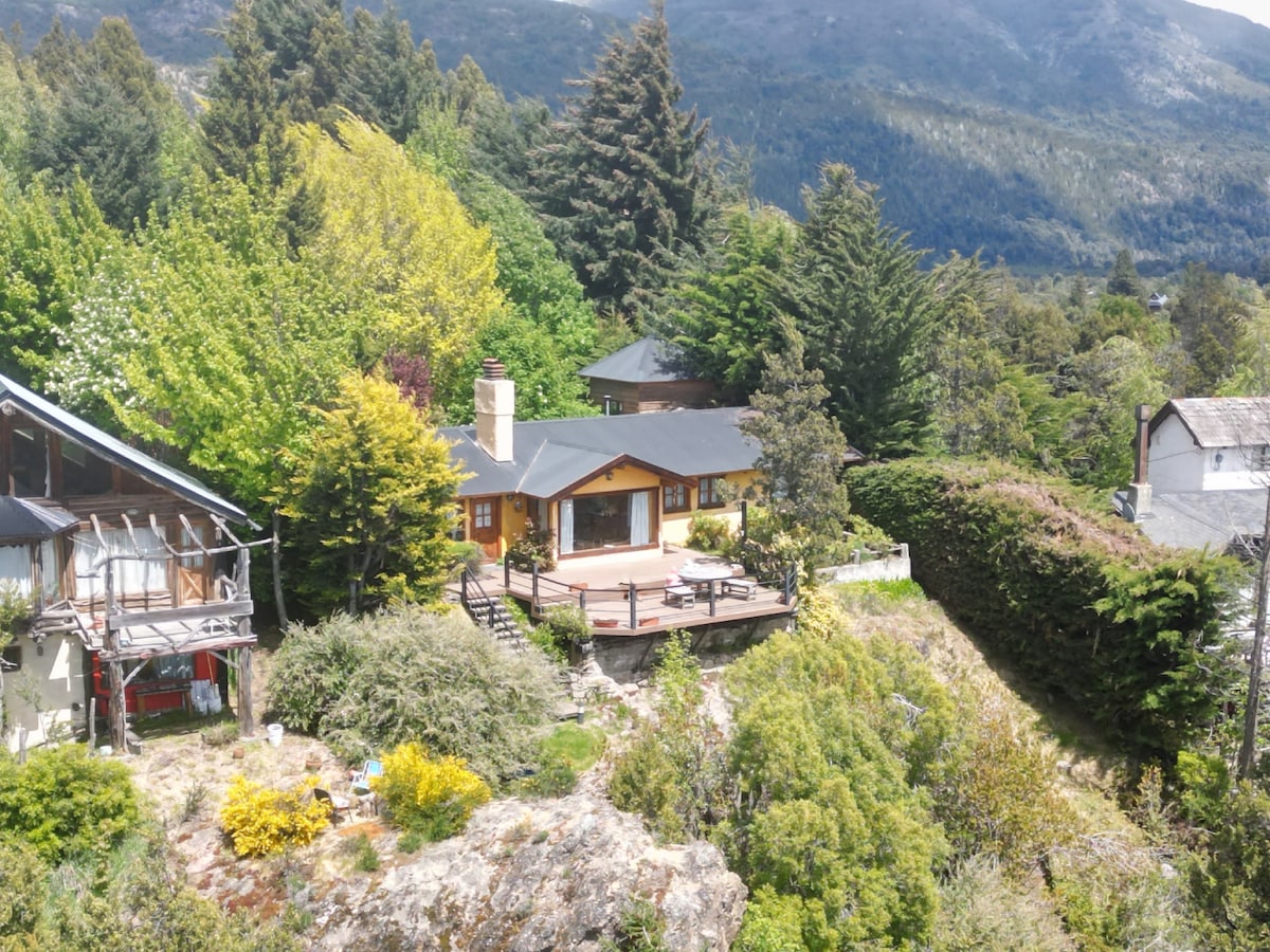 The exterior features a classic mountain house surrounded by lush greenery. A spacious deck extends from the house, providing a scenic view of the landscape. Natural terraces and vibrant plant life are visible in the garden, enhancing the outdoor experience.