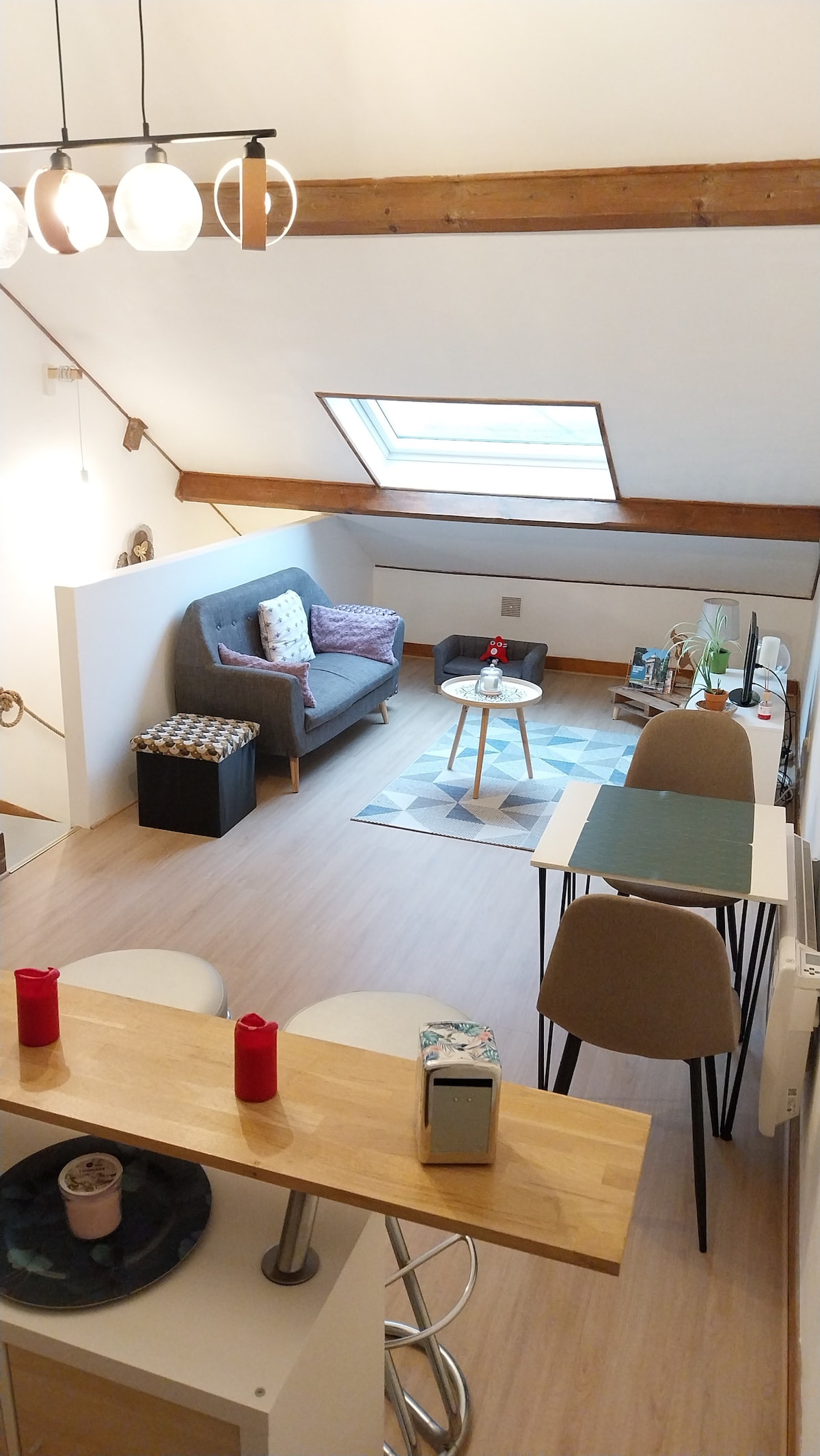 A modern living space is shown, featuring a gray sofa adorned with colorful cushions. A small coffee table sits in the center on a patterned rug, complemented by a dining area with a green table and chairs. Natural light filters in through a skylight, enhancing the interior.