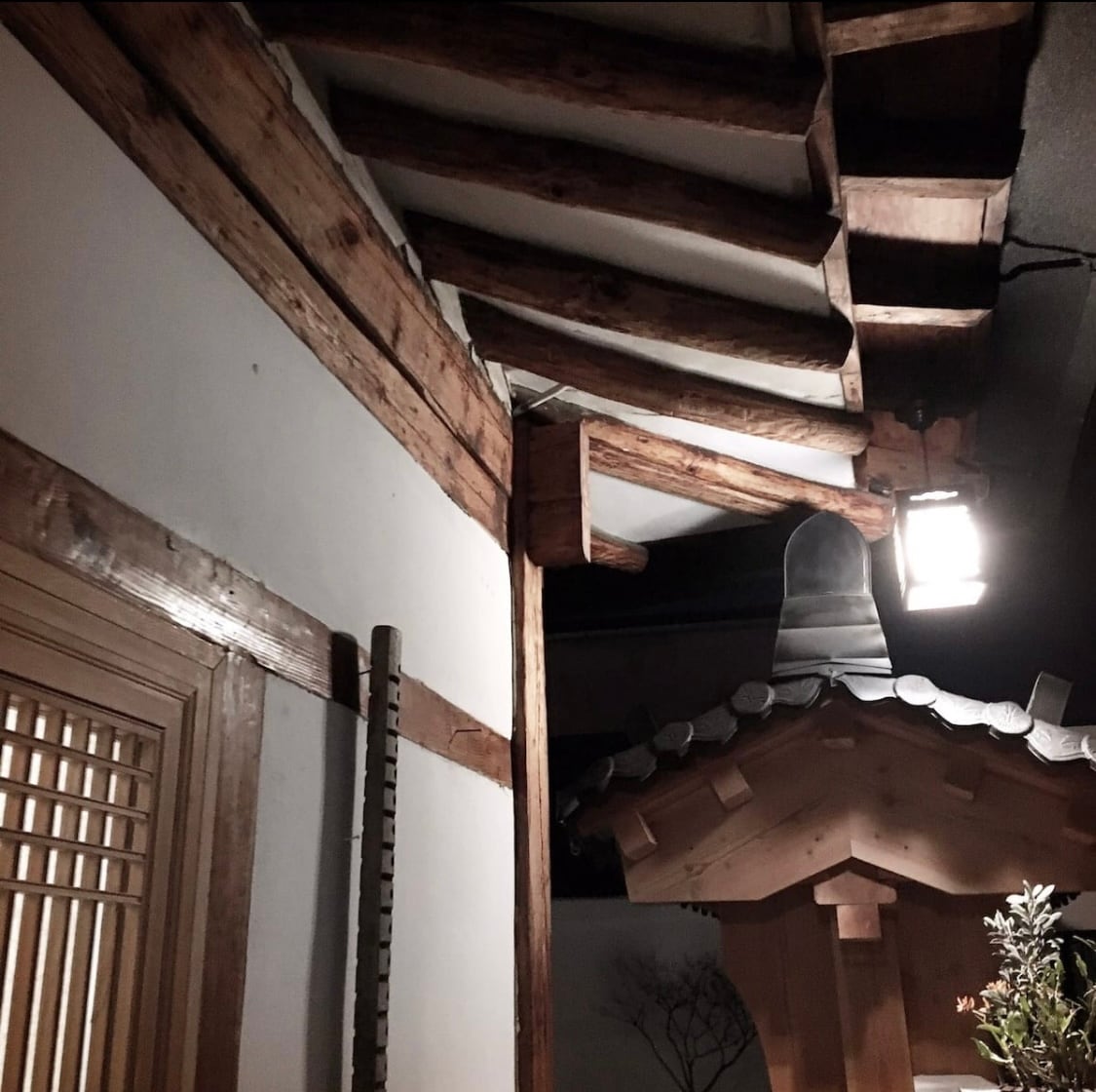 The interior of the traditional hanok showcases wooden beams with a warm finish, complementing the white walls. A softly glowing light fixture casts a gentle illumination, enhancing the architectural details. The roof features traditional tiles, contributing to the authentic aesthetic of the space.