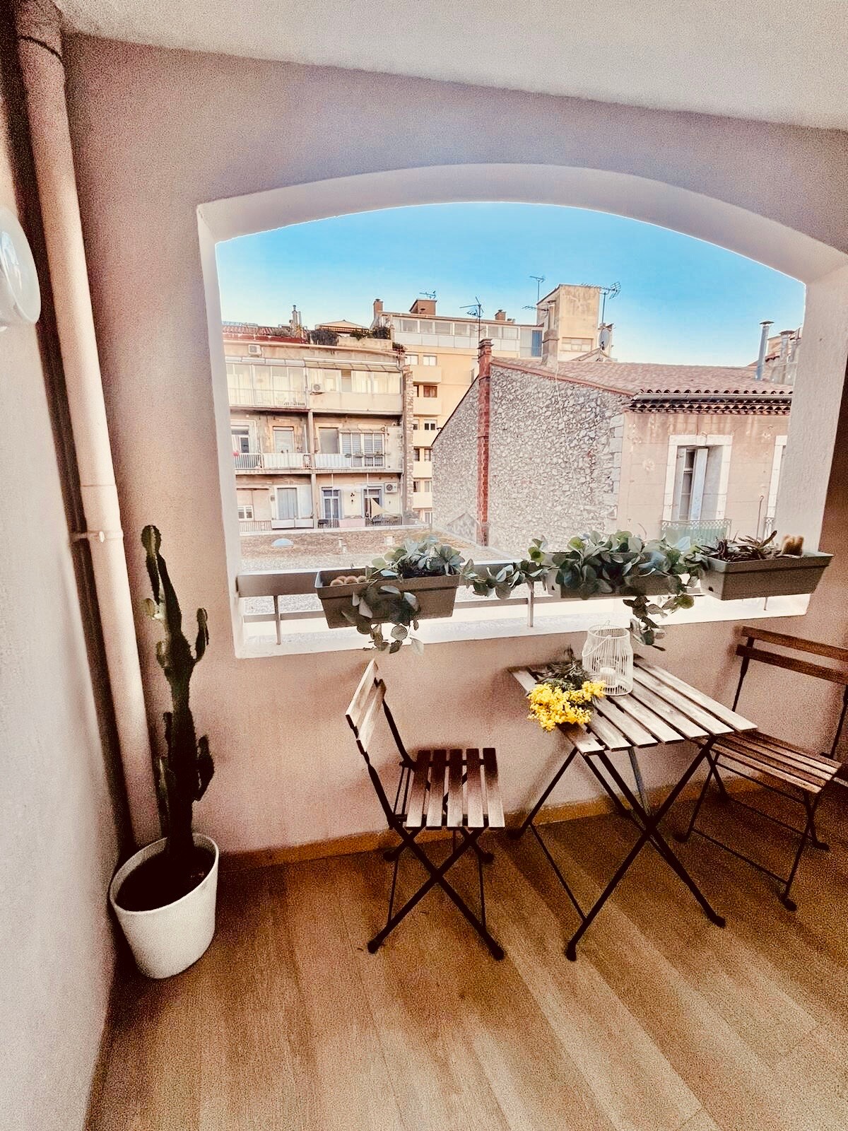 A small terrace features two wooden chairs and a folding table, adorned with fresh flowers. Potted plants are visible on the window sill, while a cactus stands in a white pot. The view showcases nearby buildings and a clear sky.