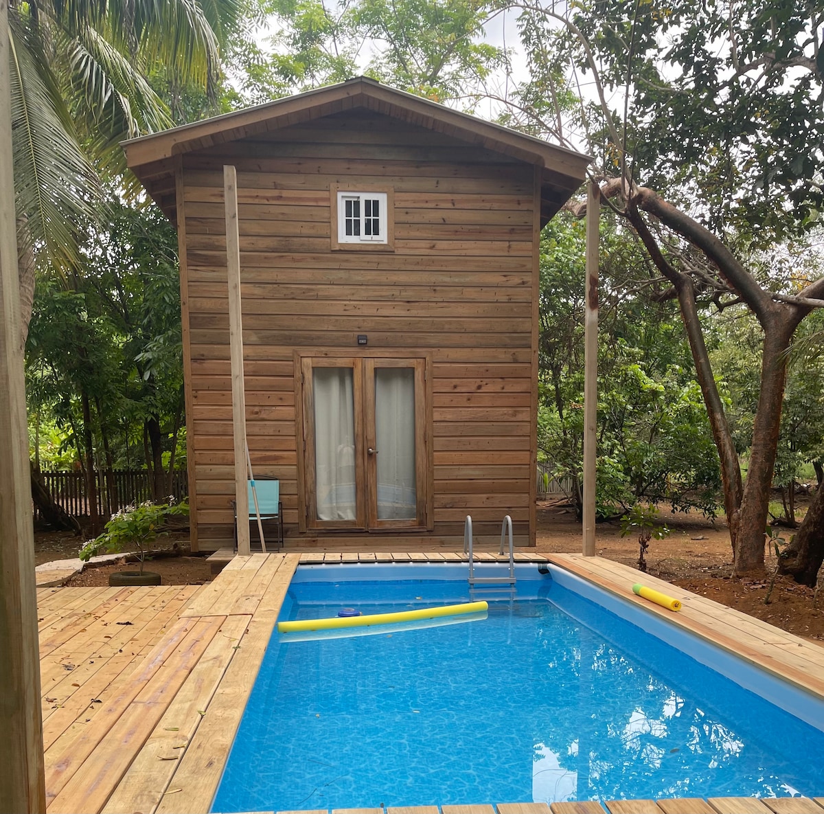 A wooden cabin features large glass doors that open to a private pool area. The pool is surrounded by a wooden deck and includes floating toys. Lush greenery is visible surrounding the property, creating a serene outdoor environment.