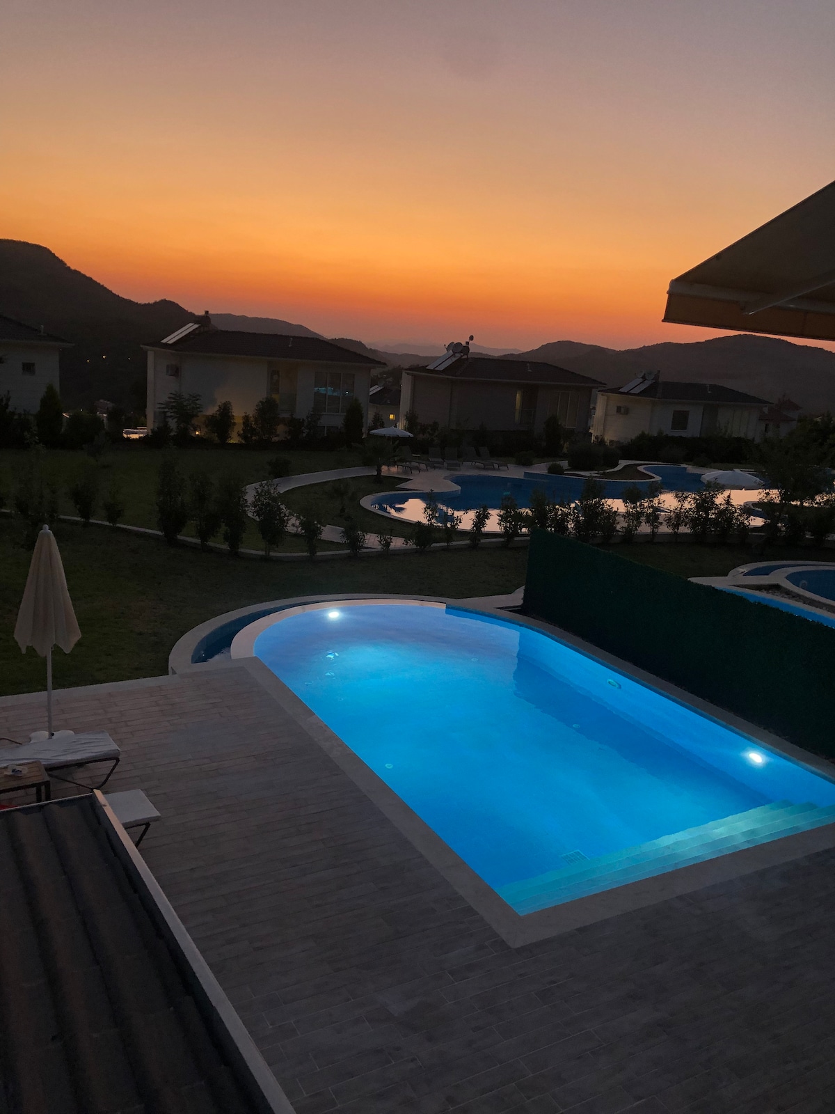 A serene evening scene reveals a glistening private pool illuminated in blue light, surrounded by well-maintained gardens. The horizon showcases a soft sunset of orange and purple hues, enhancing the tranquil atmosphere. Additional pools can be observed in the background, further complementing the setting.