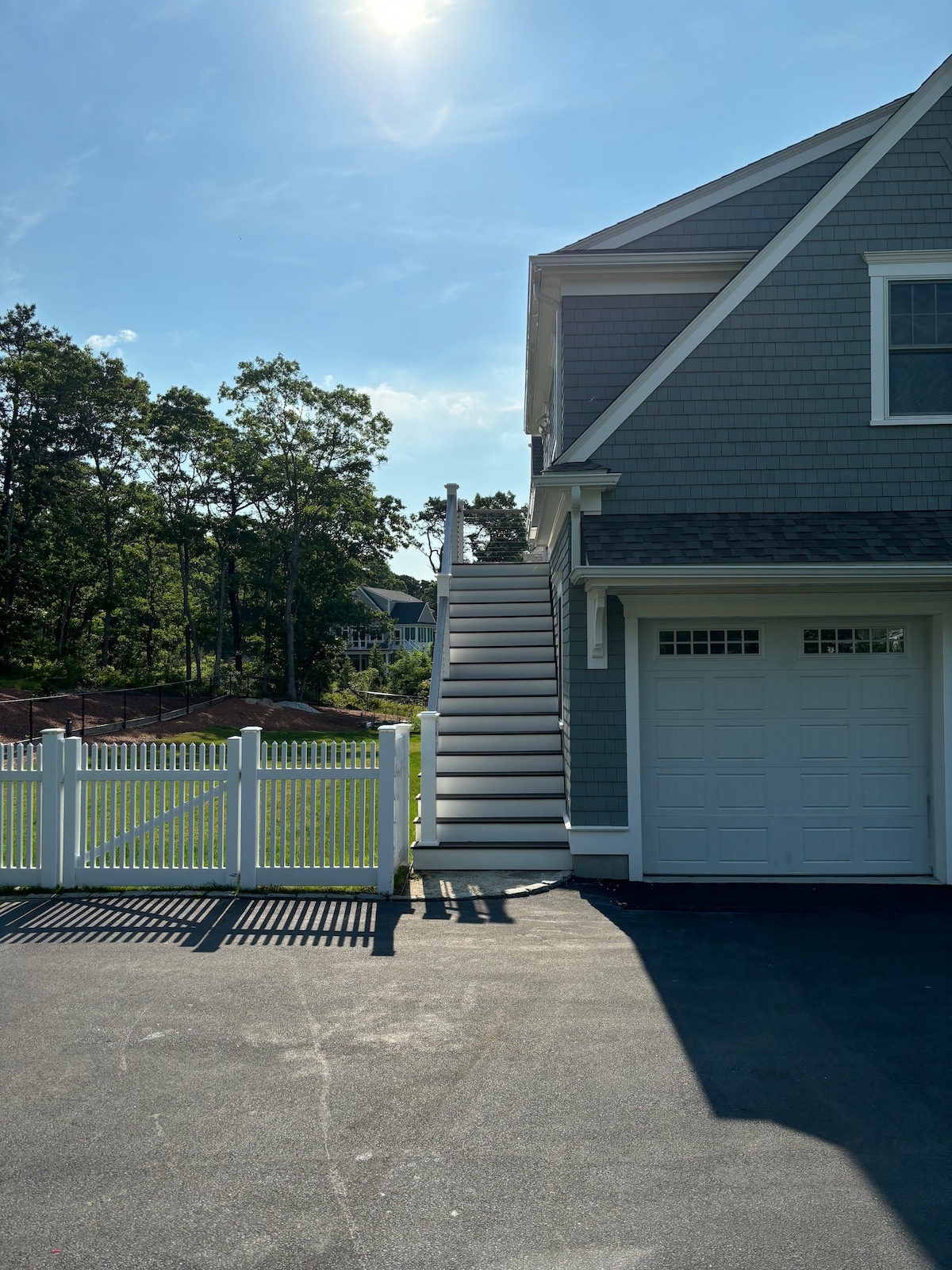 A gray exterior of a two-story structure is depicted, featuring a well-maintained staircase leading to the upper level. A white picket fence borders the property, and a sunlit driveway extends in front of a single garage door. Greenery is visible in the background.