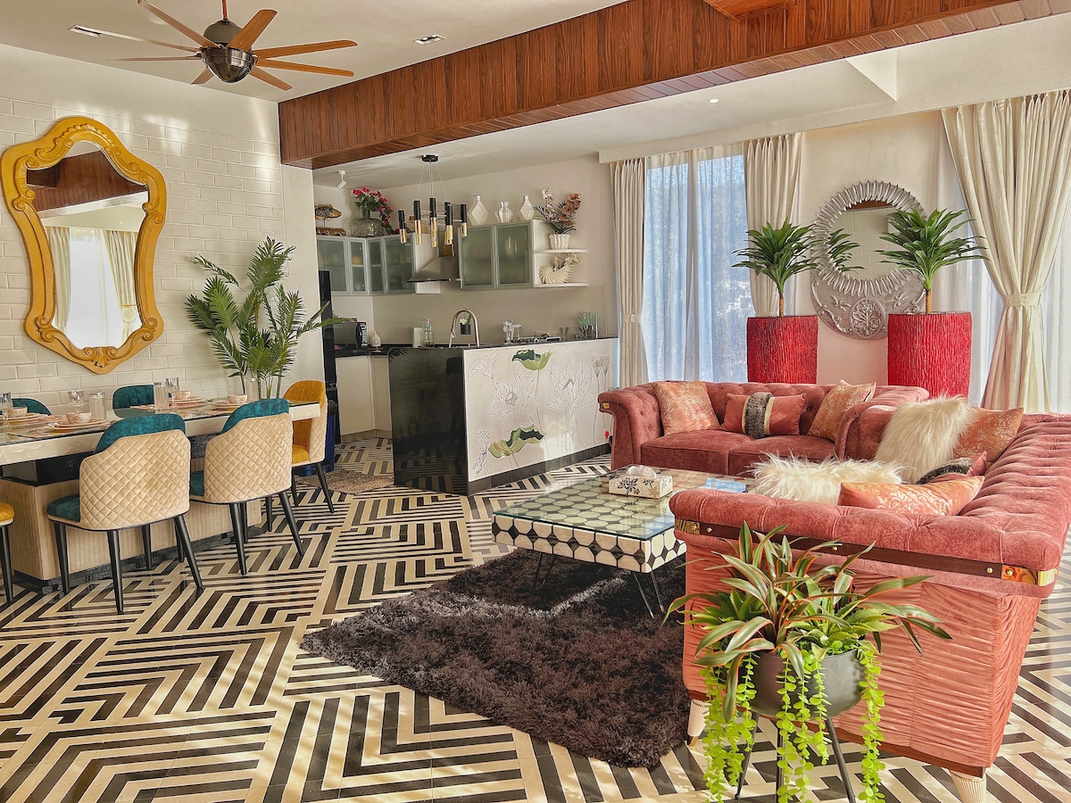 An open living area features a modern kitchen with glass cabinetry and a large dining table surrounded by colorful chairs. A plush lounge area is adorned with vibrant furniture, a textured rug, and decorative plants. Natural light pours in through the floor-to-ceiling windows, creating a bright space.