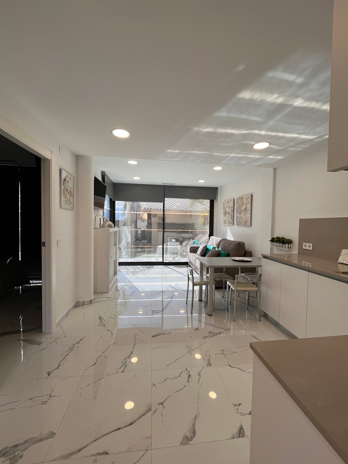 A modern kitchen area features sleek countertops and bright cabinetry. Seating is provided around a small dining table with colorful cushions. Large sliding glass doors allow natural light to flow in, connecting the interior with the outdoor space beyond. The floor is covered in glossy white tiles.