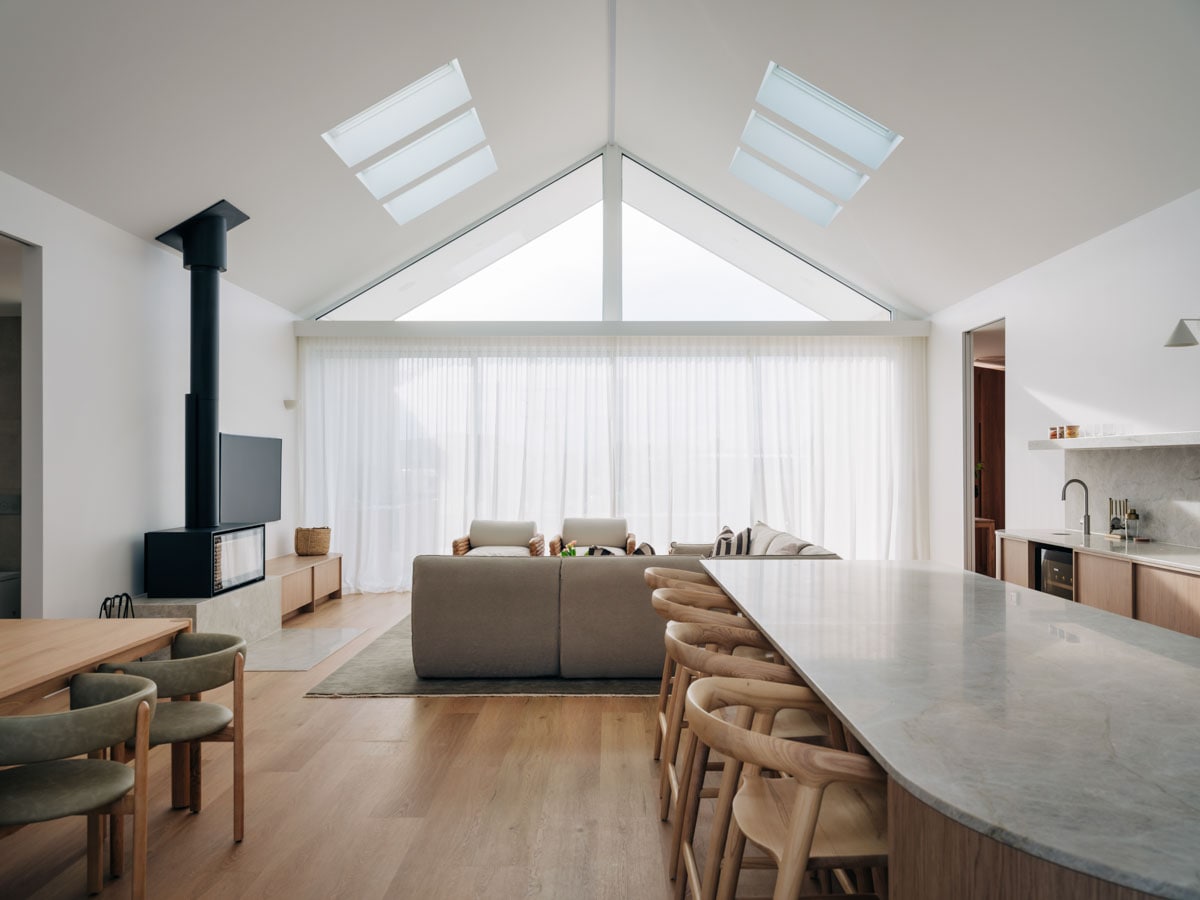 An open-plan living area features high ceilings with skylights, allowing natural light to fill the space. A comfortable sofa faces a sleek entertainment unit, while a dining table and wooden chairs are positioned nearby. A modern kitchen, complete with a stone countertop, is in view.