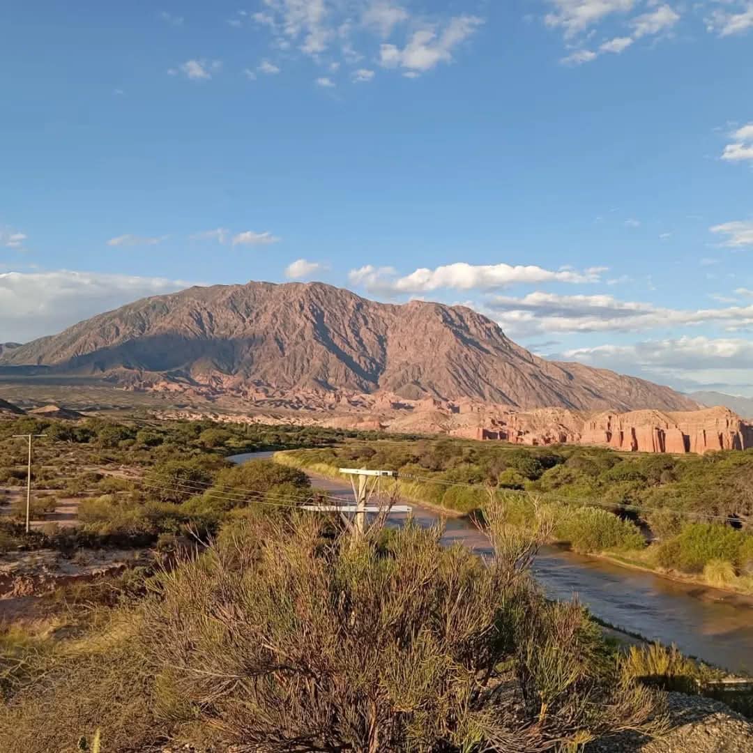 A panoramic view captures a rugged mountain landscape, where a river gently winds through lush greenery. The mountain's textured slopes contrast with the clear blue sky, dotted with soft clouds, creating a serene natural backdrop.