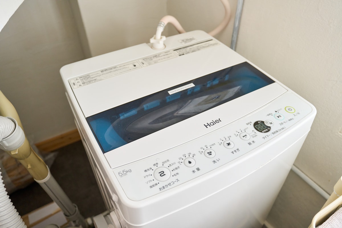 The image displays a top-loading washing machine positioned in a compact laundry area. The machine features a clear lid, control panel with various settings, and is equipped with a drainage hose. The surroundings appear tidy and organized.