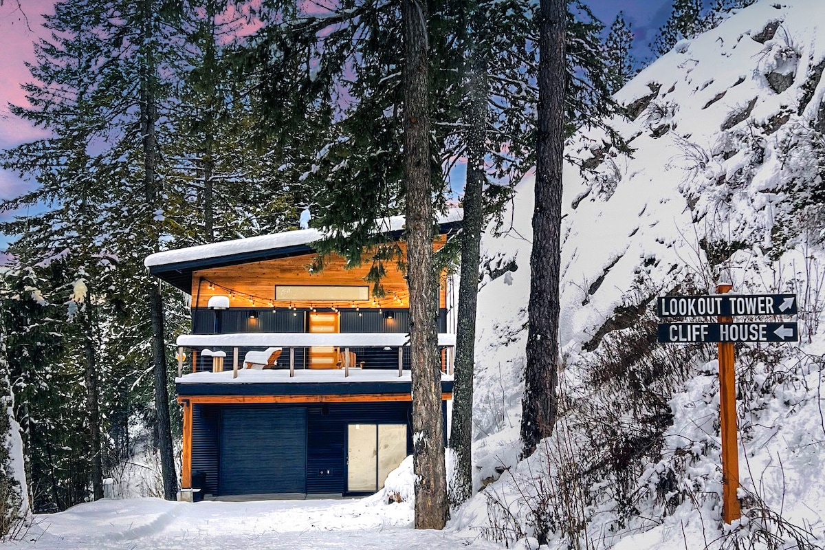 A contemporary two-story vacation rental is nestled among snowy pines. A spacious deck is visible, along with a signpost indicating directions to Lookout Tower and Cliff House. The structure features large windows, inviting natural light into the interior.