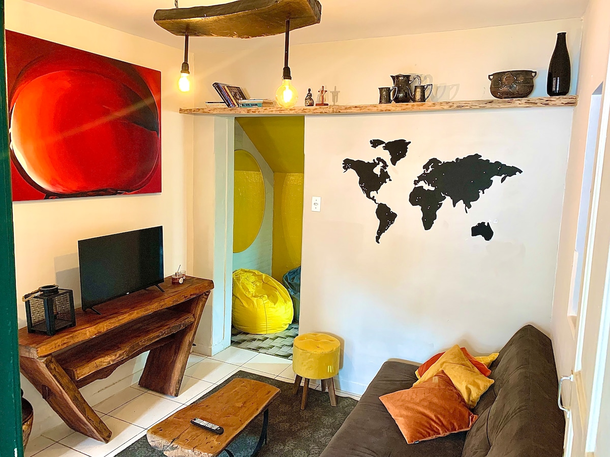 A cozy living area is showcased, featuring a comfortable sofa adorned with colorful cushions. A wooden table is positioned next to a television. The wall displays a world map, while a charming shelf holds decorative items. An inviting yellow nook is visible in the background.