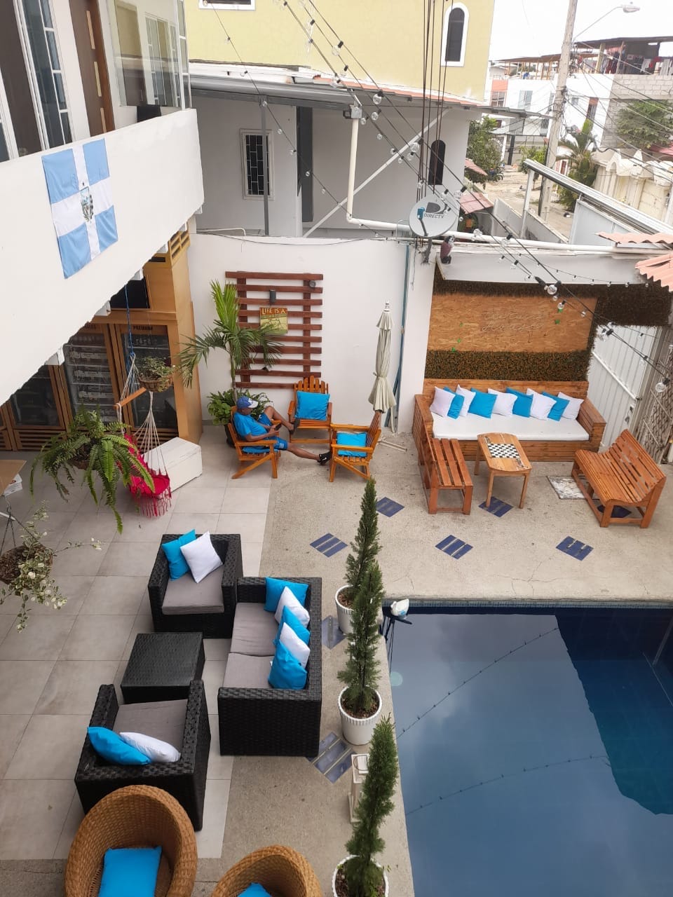 An outdoor area is presented, featuring a swimming pool surrounded by lounge seating. A mix of wicker chairs and wooden benches creates a casual gathering space. Colorful cushions add vibrancy, while tall potted plants provide greenery around the pool area. Decorative string lights are strung overhead.