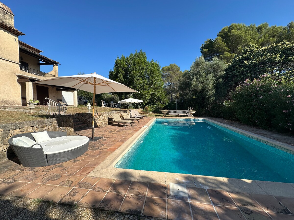 An inviting outdoor space features a rectangular pool surrounded by tiled flooring. Sun loungers and umbrellas provide shaded seating areas. Lush greenery and trees create a tranquil backdrop, while a stone wall adds to the privacy of the setting.