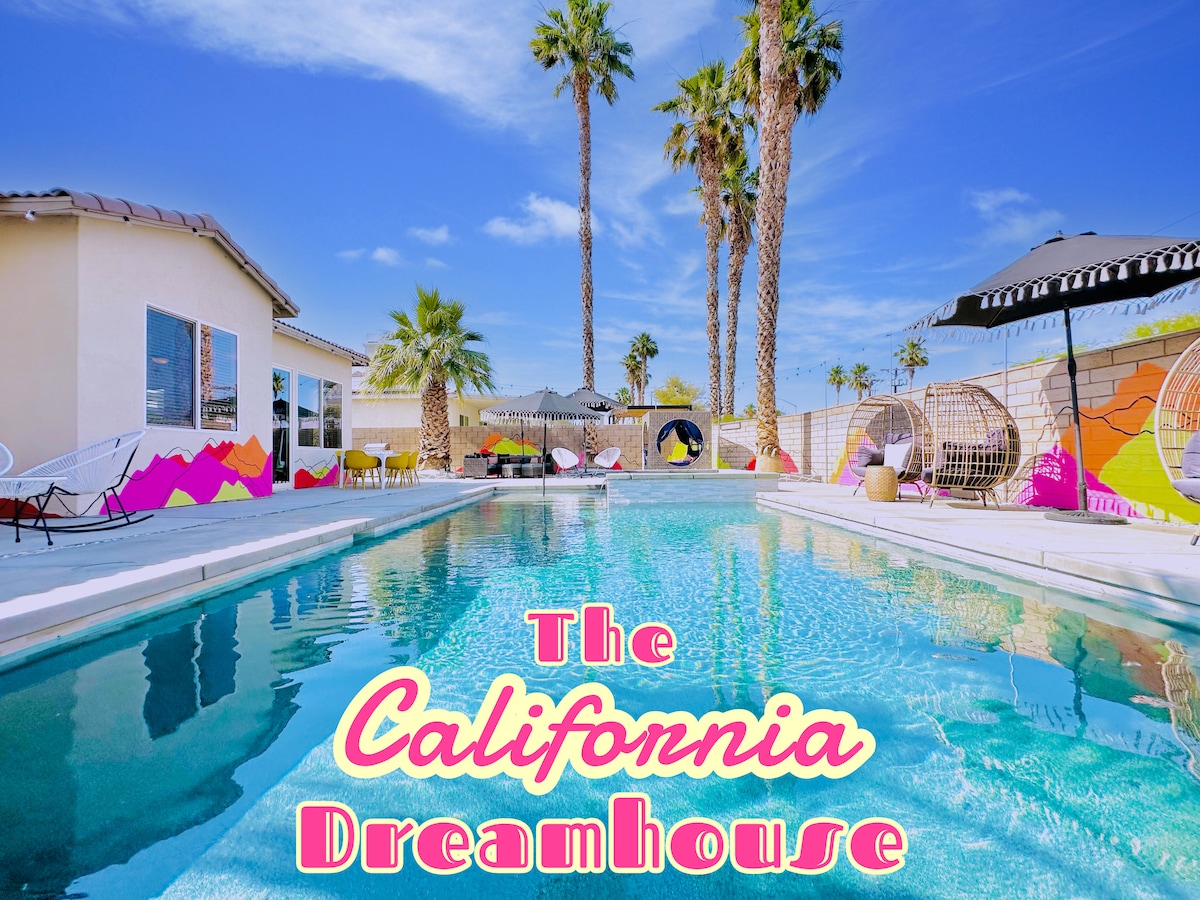 A serene view of the pebble-tech saltwater pool is framed by tall palm trees. Colorful custom murals and inviting lounge chairs are visible along the poolside, creating a vibrant atmosphere. The spacious patio provides ample area for relaxation and enjoyment under a clear blue sky.