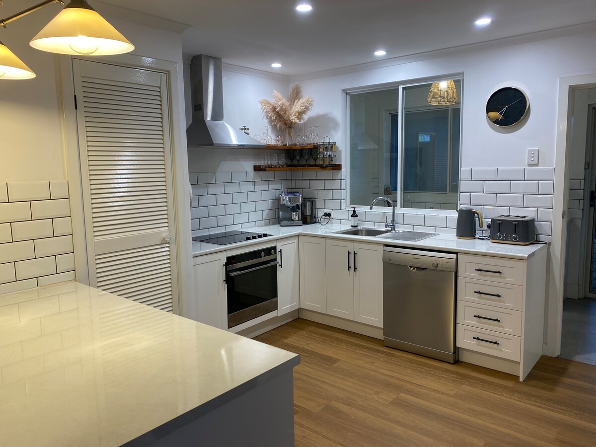 A modern kitchen features polished countertops and white cabinetry, with stainless steel appliances including an oven and dishwasher. Natural light fills the space through a large window above the sink, complementing the tiled backsplash and minimalist decor.