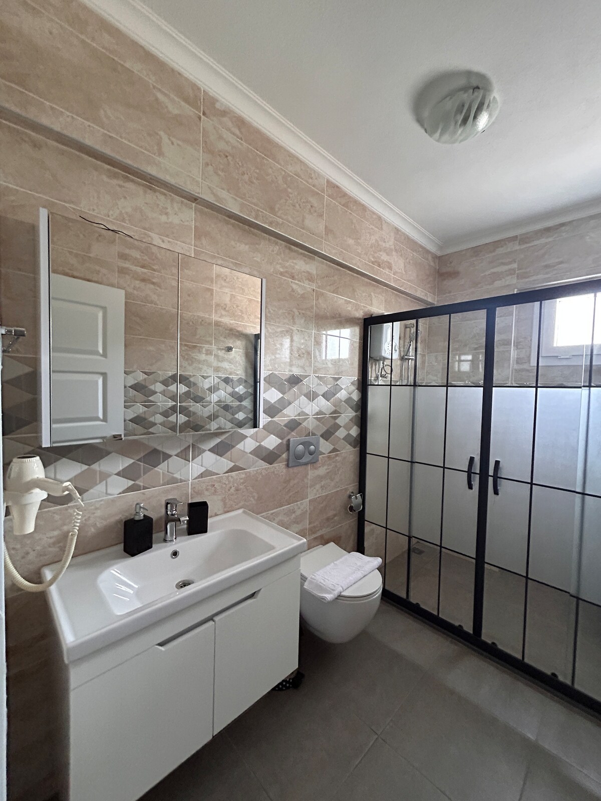 A modern bathroom features beige tiled walls and a glass-enclosed shower. A white vanity is equipped with a mirror and sink, while a compact toilet is positioned nearby. Fresh towels are neatly arranged on the vanity, enhancing the clean and organized appearance.