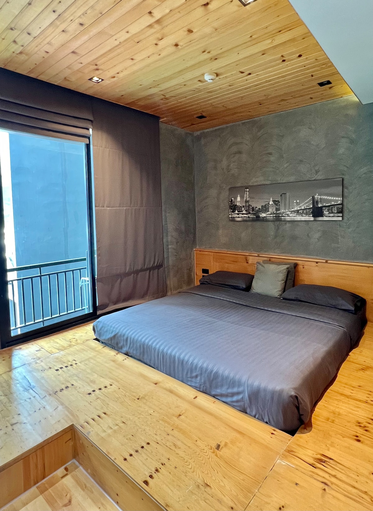 A spacious sleeping area features a wooden platform bed with a soft, dark grey blanket. Natural light enters through a large window covered by elegant dark curtains. Simple artwork adorns the wall, and wooden ceiling beams enhance the room's design.