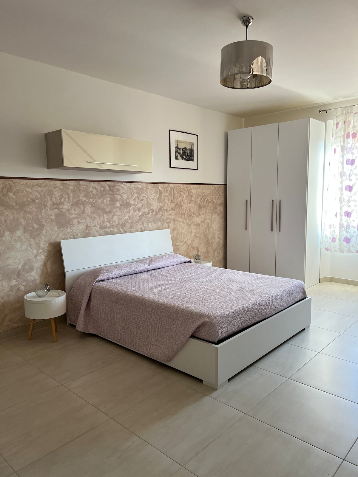 A simple bedroom features a spacious bed covered with a light lavender blanket. A nightstand stands next to the bed, and a wardrobe is positioned against one wall. Natural light enters through a window adorned with sheer floral curtains.