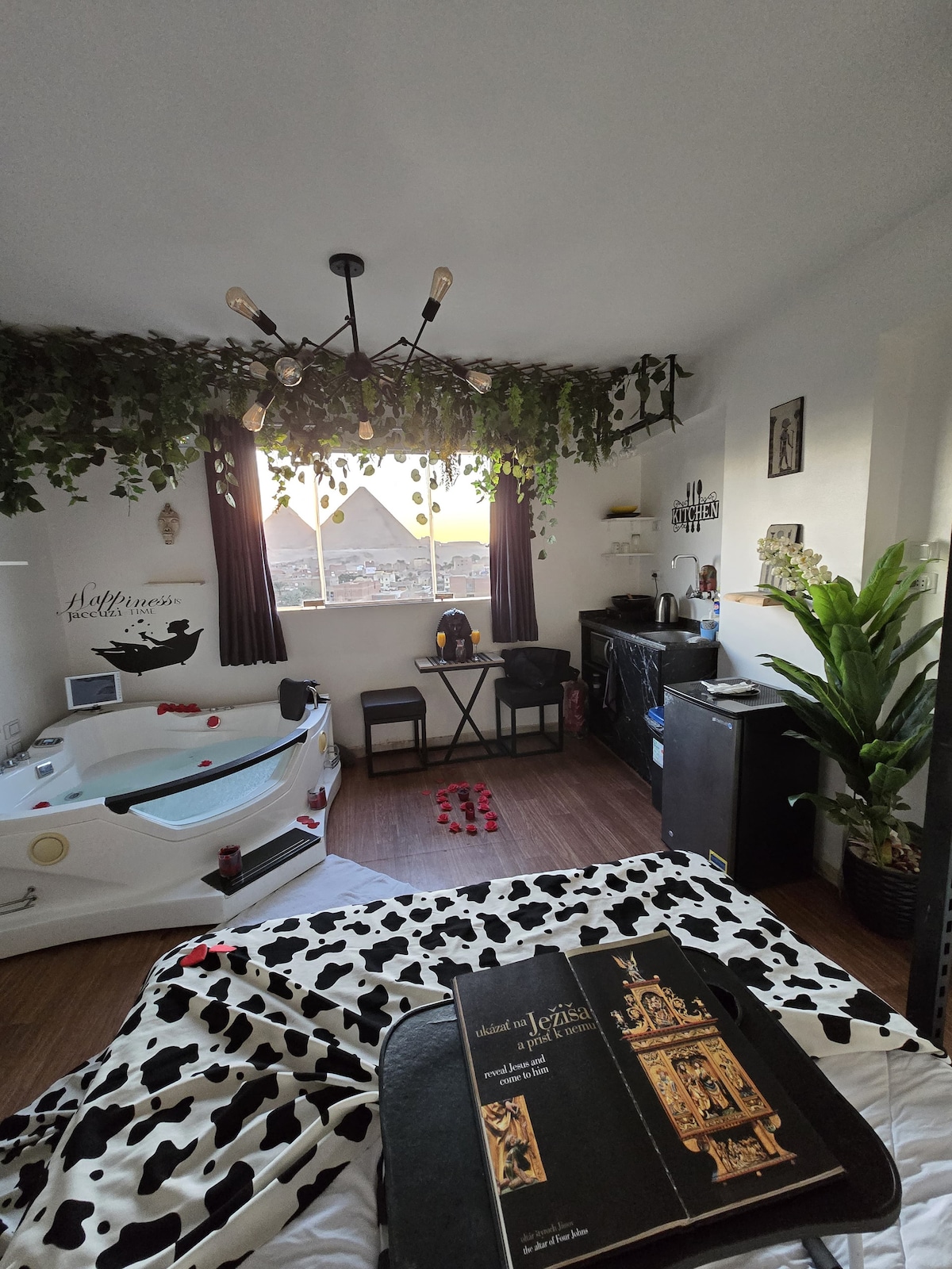 A cozy room features a jacuzzi adorned with rose petals, a small dining table, and a compact kitchenette. Large windows allow natural light to enter, framed by dark curtains and lush greenery. A stylish book rests on the bed, enhancing the inviting decor.