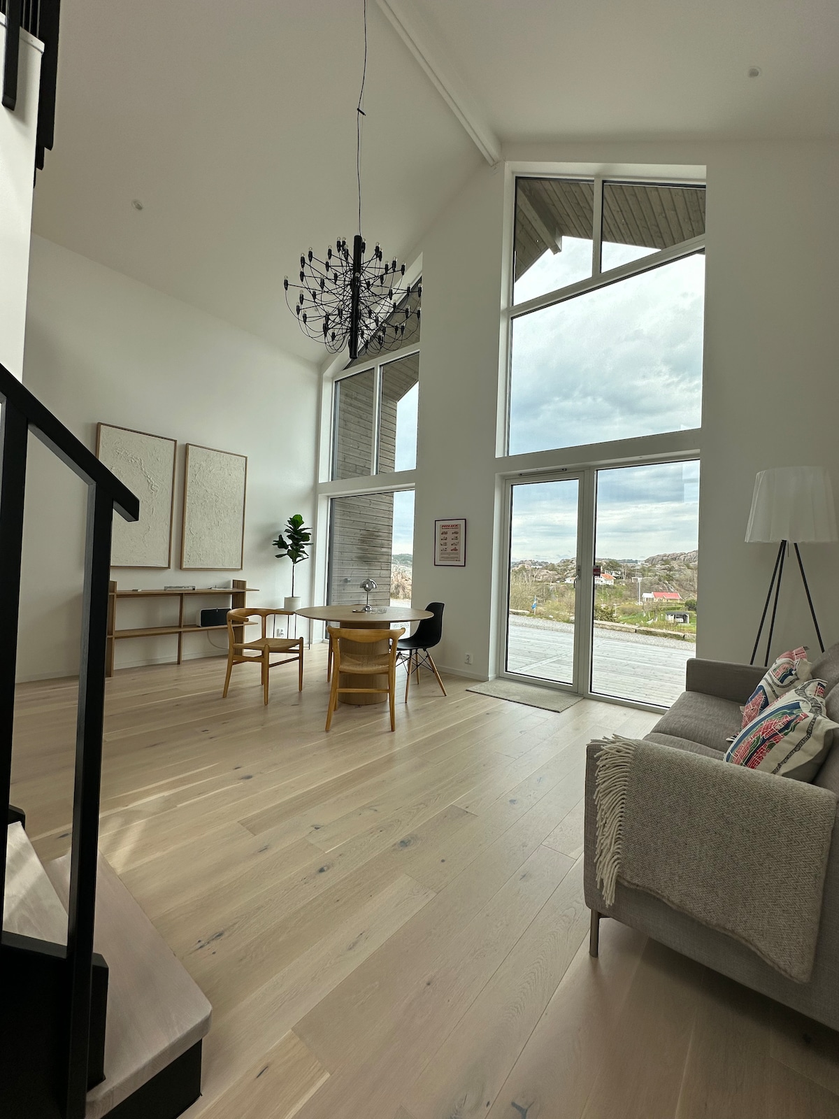 A bright living area is defined by tall windows allowing ample natural light to fill the space. A modern chandelier hangs from the high ceiling. A cozy sofa is paired with wooden dining chairs and a table, accompanied by subtle decor elements against a light wall.