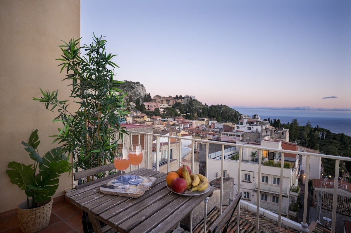 A small balcony is featured with a wooden table set for two, holding drinks and a plate of fruit. Lush green plants are positioned nearby, while a panoramic view of Taormina and the sea is visible in the background, set against a soft evening sky.