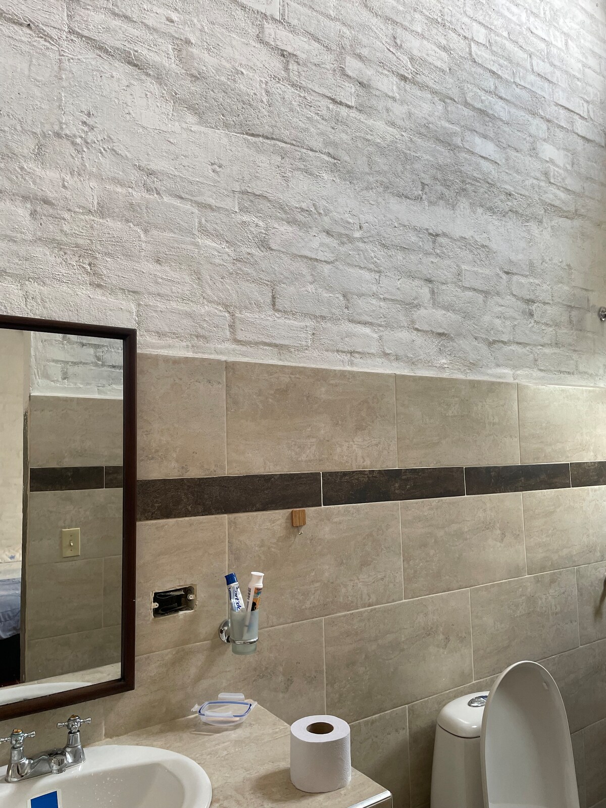 A modern bathroom features a light brick wall alongside elegant stone tiles. A mirror is mounted above a sleek sink, which is paired with a stylish faucet. A toilet is positioned nearby, and a roll of toilet paper is neatly arranged on the countertop.