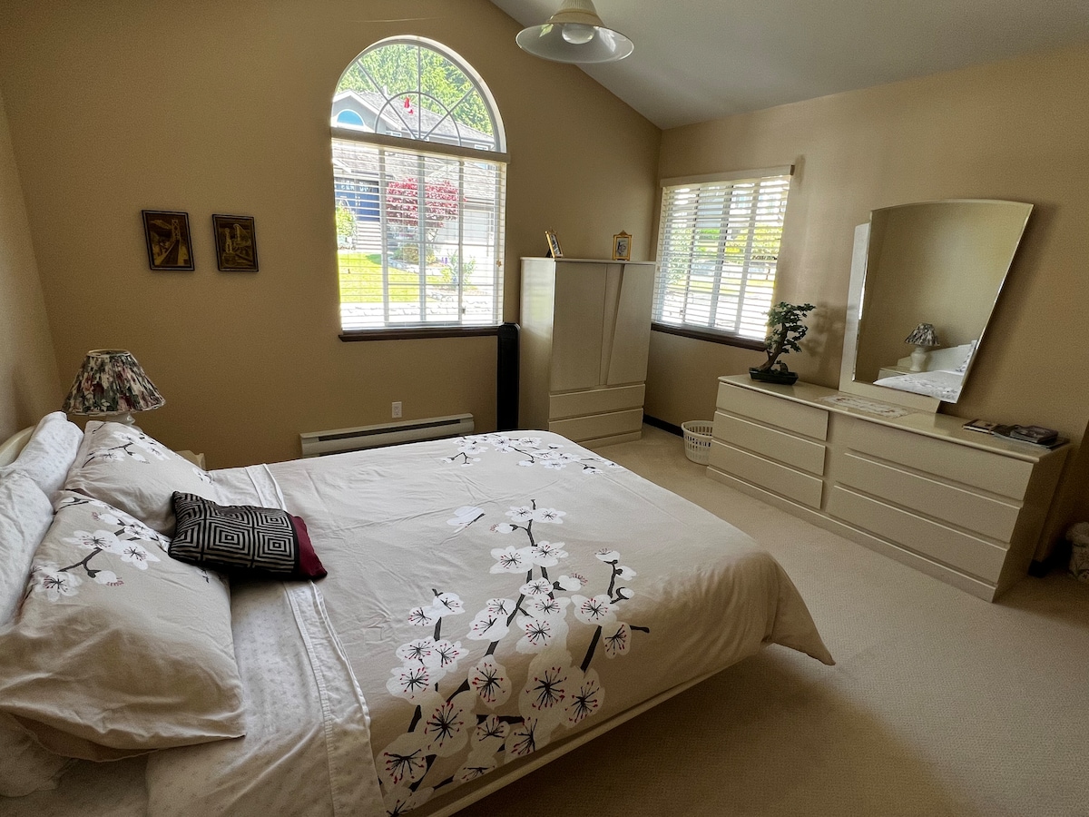 A spacious bedroom features a large bed adorned with floral bedding and accent pillows. Natural light filters through an arched window, illuminating the room. A mirror and dresser are positioned against one wall, complemented by a small decorative lamp and a leafy plant.