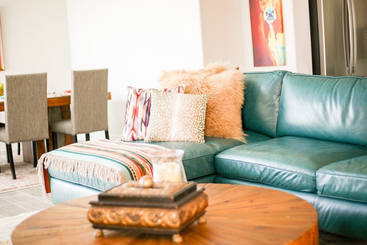 A spacious living area features a comfortable teal sectional sofa adorned with patterned pillows and a soft throw blanket. A round coffee table sits in front of the sofa, topped with a decorative bowl. In the background, a dining area is visible with a wooden table and upholstered chairs.