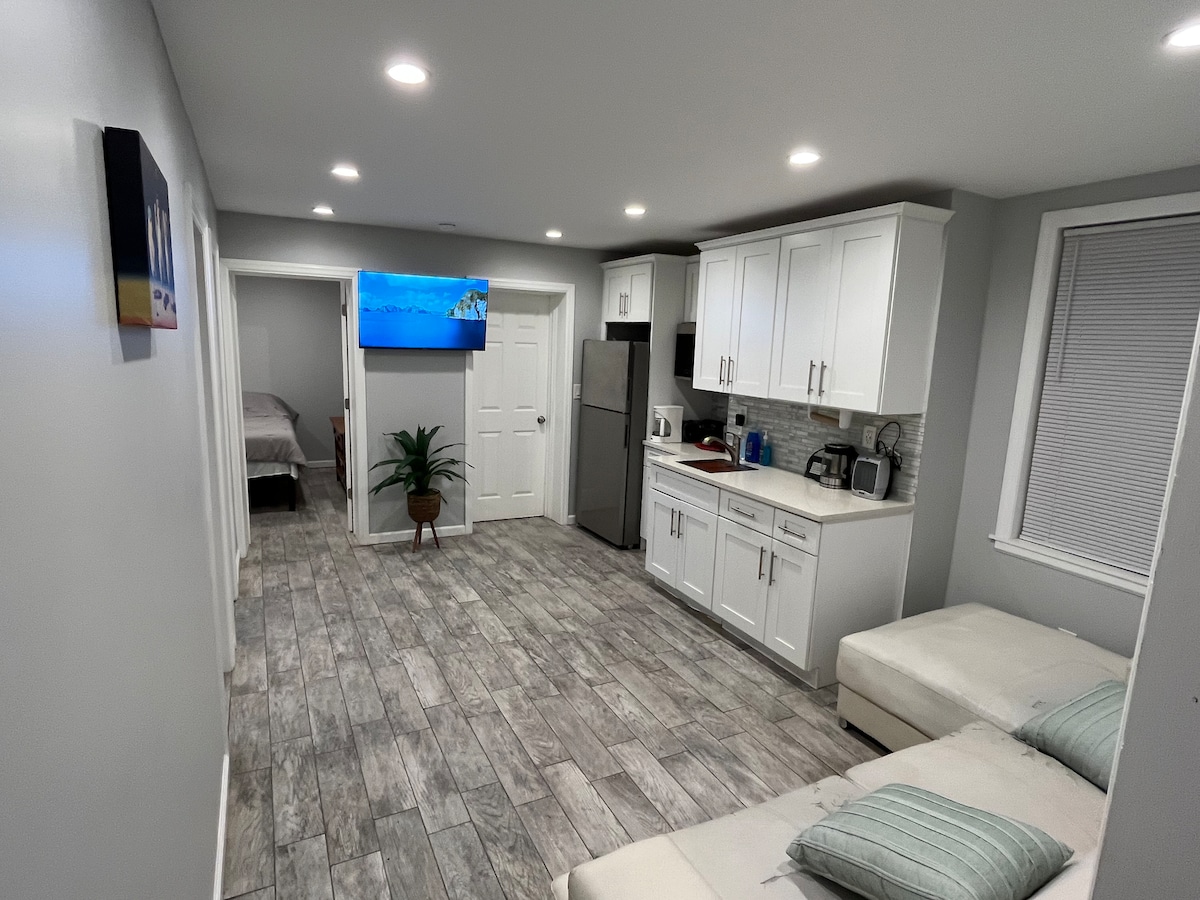 A modern living space features light grey walls and warm-toned flooring. A kitchenette with white cabinets is visible on one side, along with essential appliances. A small seating area is positioned nearby, and a wall-mounted television adds to the atmosphere. Light enters through a bright window.