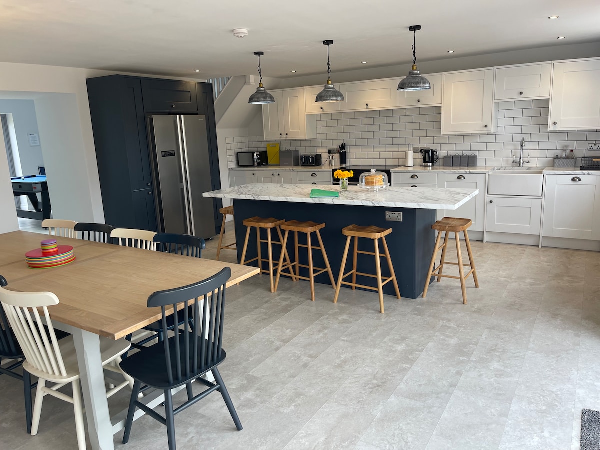 A spacious kitchen diner is equipped with modern appliances, including a large refrigerator and several countertop gadgets. A central island with bar stools provides seating, while a dining table surrounded by various chairs accommodates group meals. The light-coloured cabinetry is complemented by white tiled backsplash.