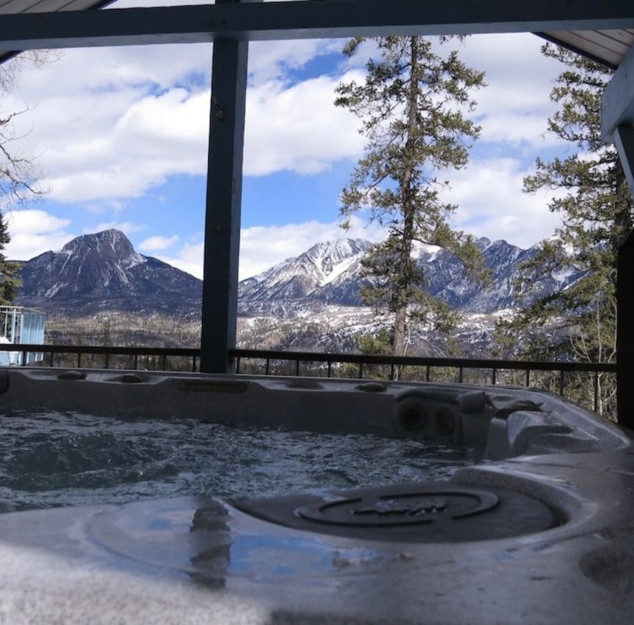 A hot tub is positioned under a covered area, revealing panoramic views of snow-capped mountains in the distance. The surrounding trees frame the landscape, and the sky displays scattered clouds, enhancing the serene outdoor experience.