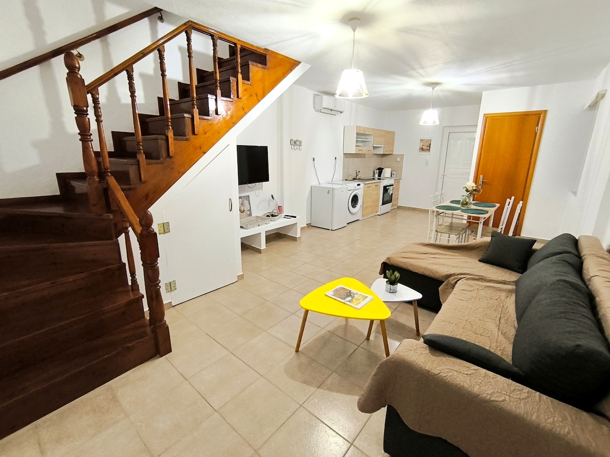 An inviting living space is depicted, featuring a comfortable sofa oriented towards a mounted television. A dining table with chairs is visible in the background, adjacent to a well-equipped kitchen. A staircase leads to the upper level, while light fixtures illuminate the room.