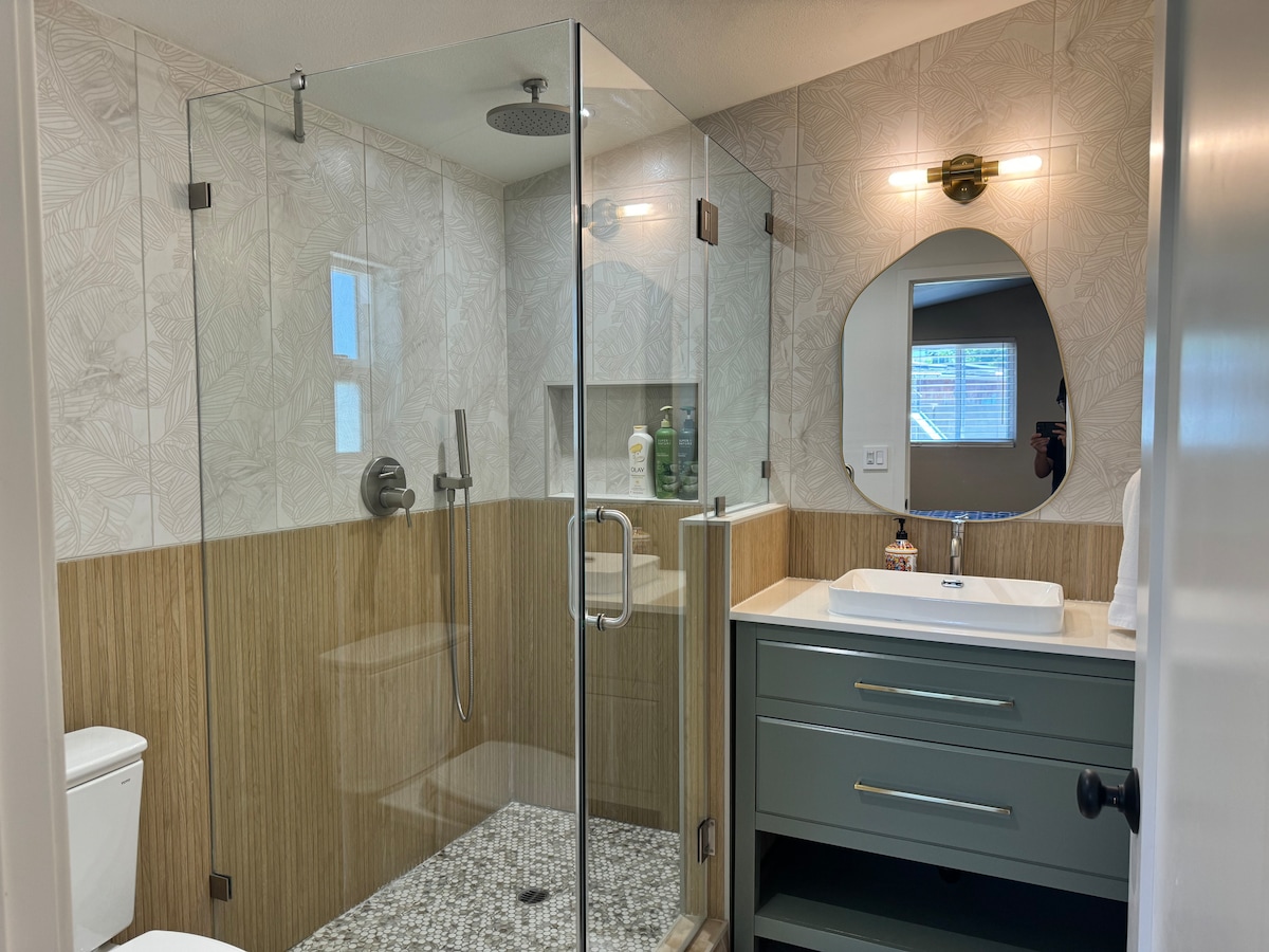 A modern bathroom features a walk-in shower with a glass enclosure and rainfall showerhead. A stylish sink sits atop a grey vanity, complemented by light wood paneling and decorative wall tiles. Natural light filters in through a window, providing a bright and airy ambiance.