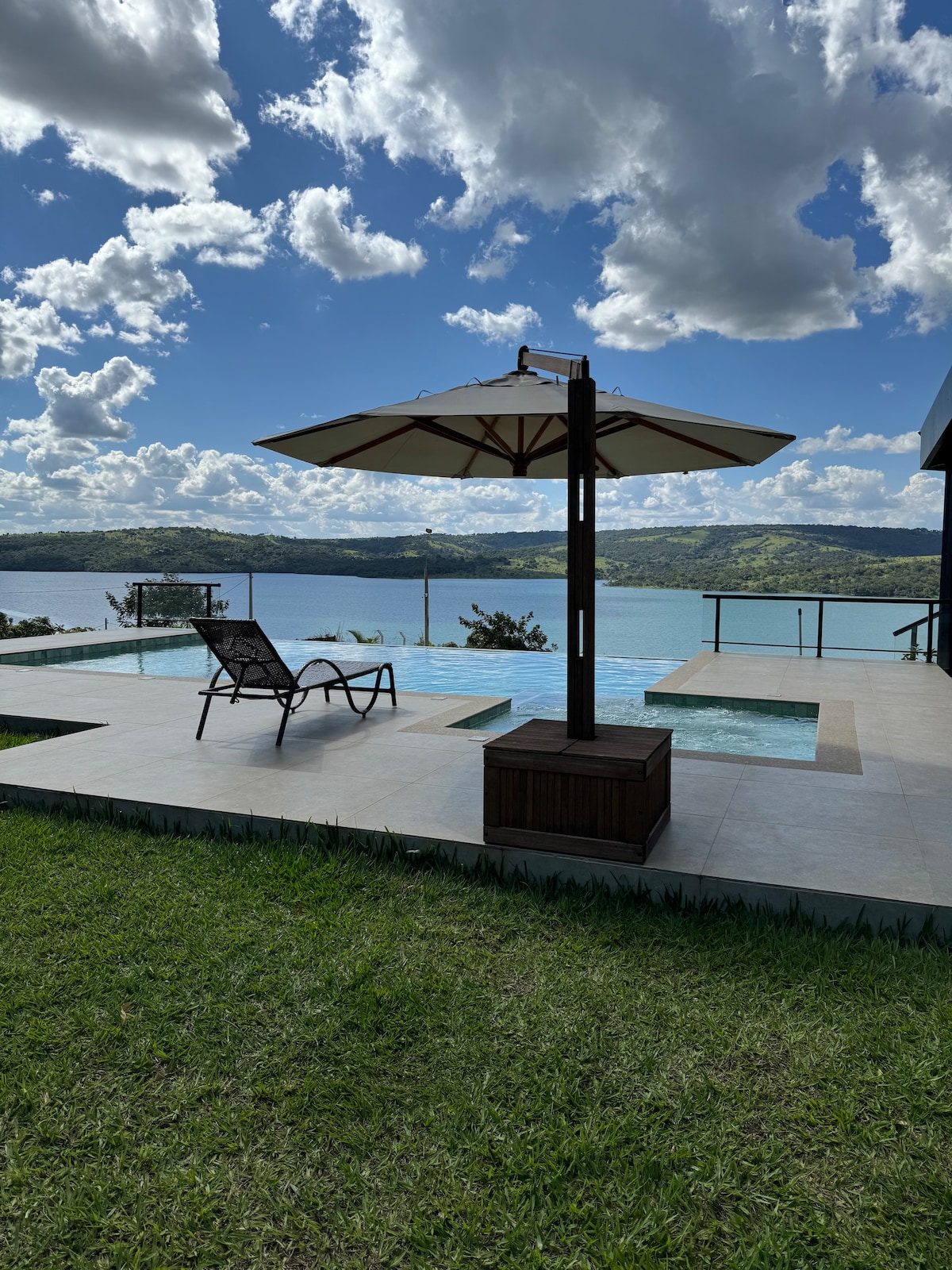 An outdoor space features a sunbed positioned under a large umbrella, overlooking a serene lake. The infinity pool extends toward the water, complemented by a stone patio and lush green lawn. Soft clouds float above, highlighting the natural beauty of the surroundings.