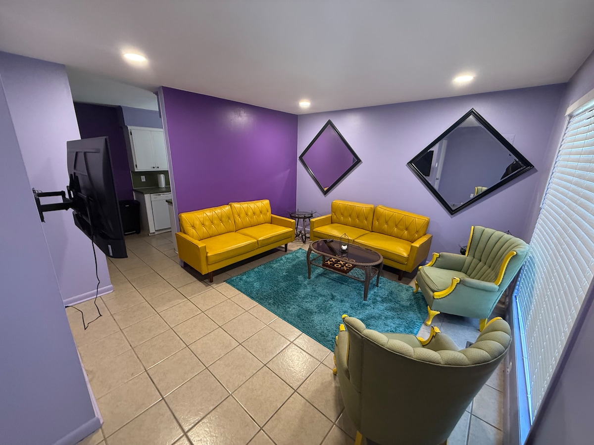 A vibrant living area showcases yellow sofas and a green accent chair, arranged around a wooden coffee table. Large mirrors adorn the walls, reflecting the space's bold purple tones. A blue area rug adds a pop of color against the tiled floor, with a television mounted nearby.