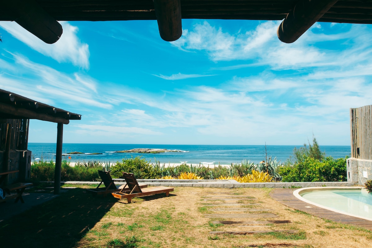 A scenic view showcases an outdoor area featuring a grassy space with wooden lounge chairs positioned for relaxation. The tranquil ocean stretches into the distance under a bright blue sky, with lush greenery framing the landscape.