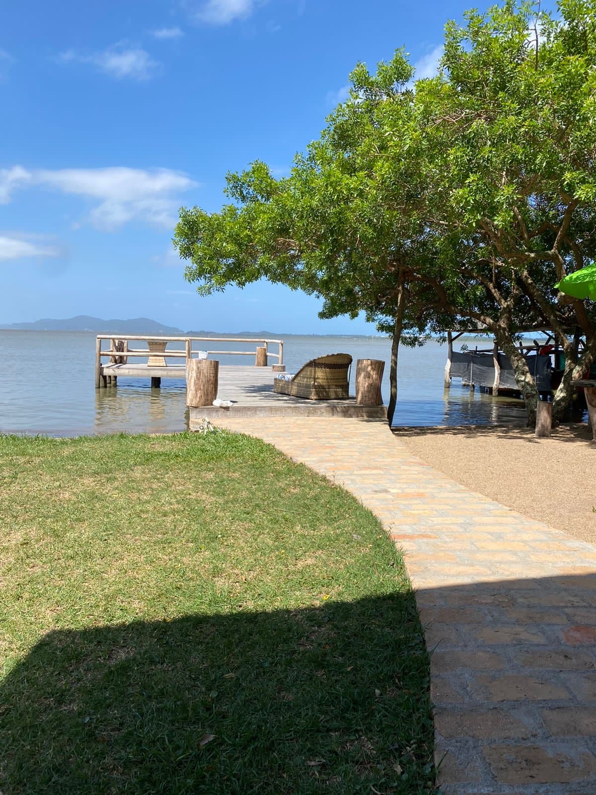 A stone pathway leads to a wooden dock adjacent to the water, framed by lush greenery. The calm lake reflects the clear blue sky, while a boat is visible nearby. The scene exudes a sense of tranquility and connection to nature.