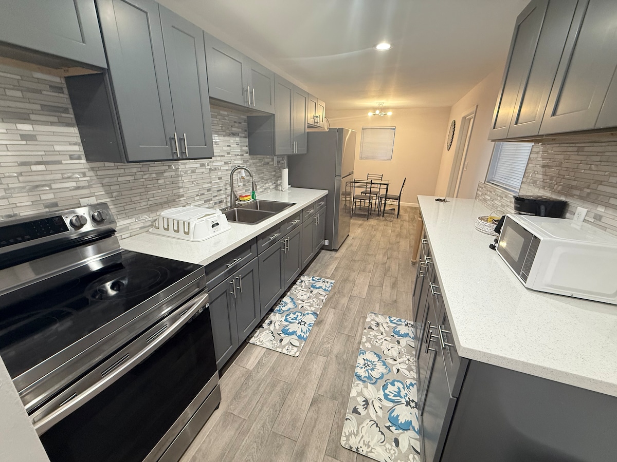 A modern kitchen features dark gray cabinetry and a spacious white countertop. Stainless steel appliances include an oven and microwave. Light gray tile flooring is complemented by decorative mats. A dining area with a table and chairs is visible in the background, with natural light coming through a window.